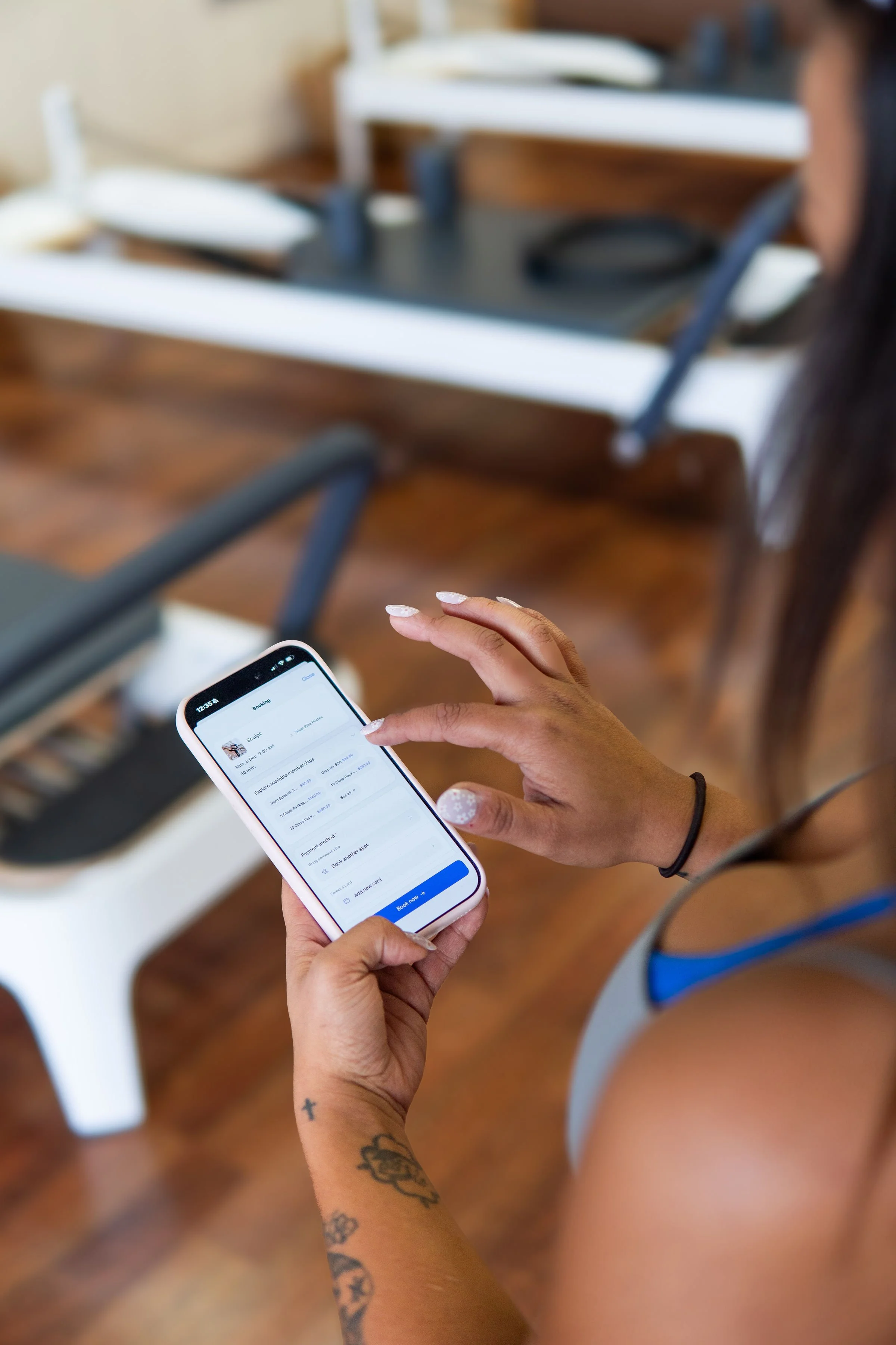 A woman with tattoos on her arm is using a smartphone to book a flight, with airport chairs and a wooden floor in the background.
