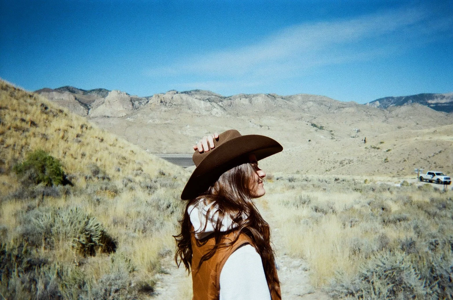 A woman standing in a desert landscape wearing a brown cowboy hat and a brown vest, with mountains in the background and a blue sky overhead.