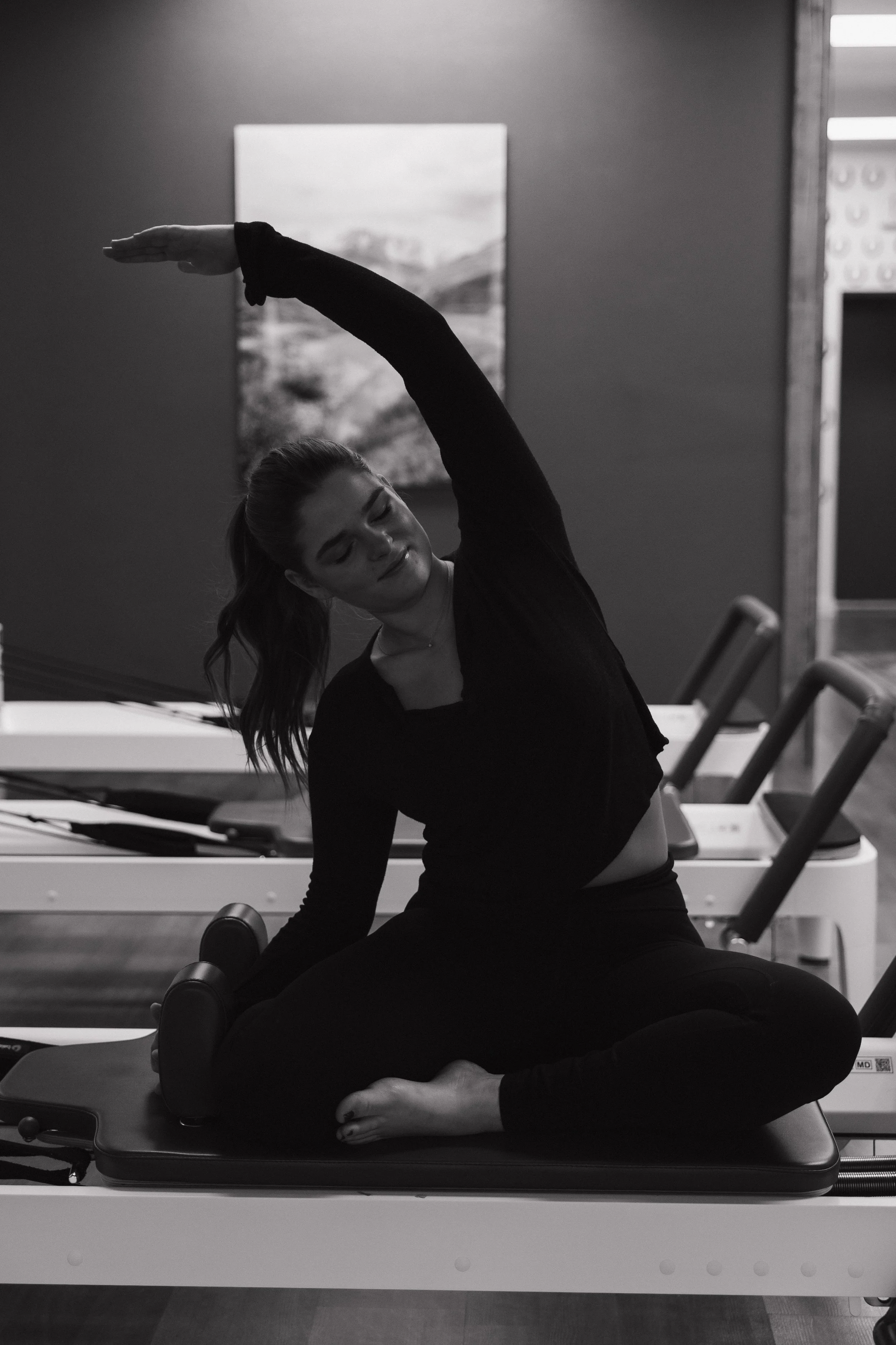 A woman practicing yoga on a pilates reformer machine, sitting cross-legged and stretching with one arm raised above her head in a fitness studio.