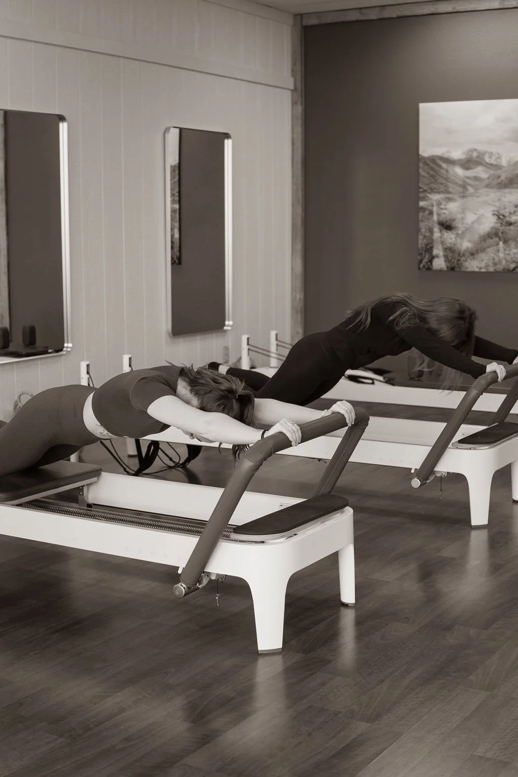 Two women are doing Pilates on reformer machines in a fitness studio, both bending forward with arms extended and heads lowered.