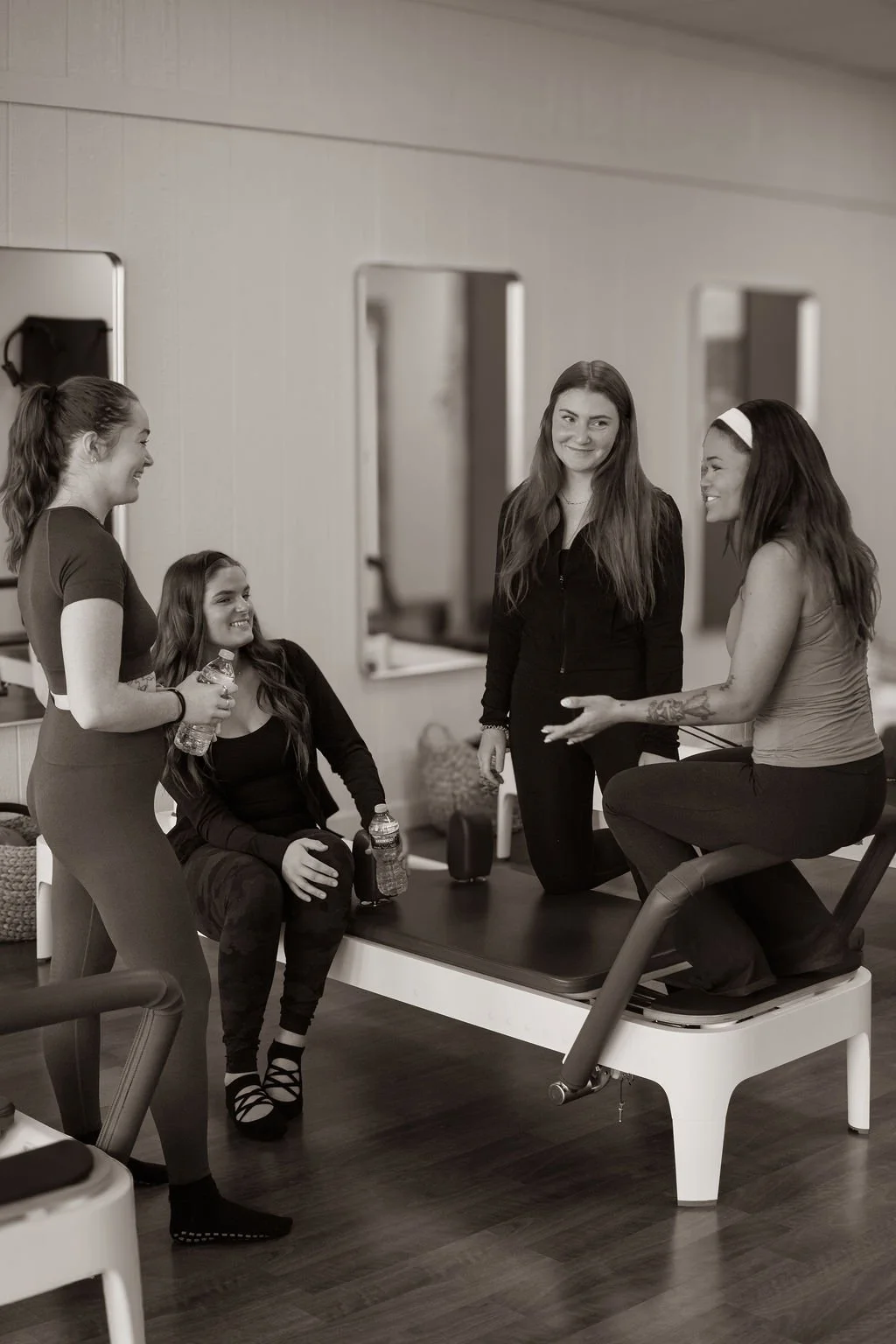Four young women having a conversation in a fitness or physical therapy setting, with one sitting on a Pilates reformer and the others standing around, holding water bottles.