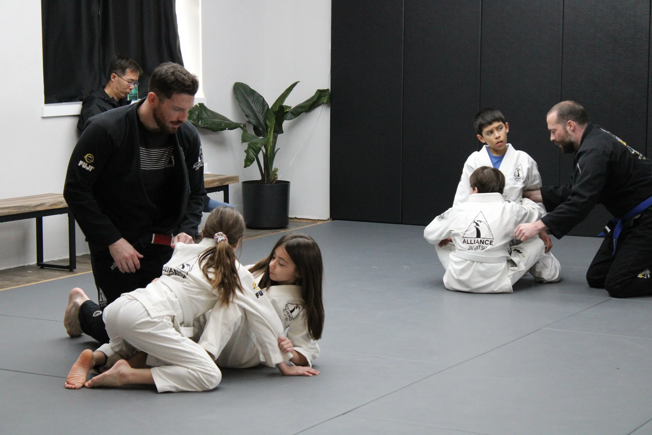 Children practicing Brazilian Jiu-Jitsu wearing white gis with instructor in black gi, engaging in ground techniques in a martial arts dojo.