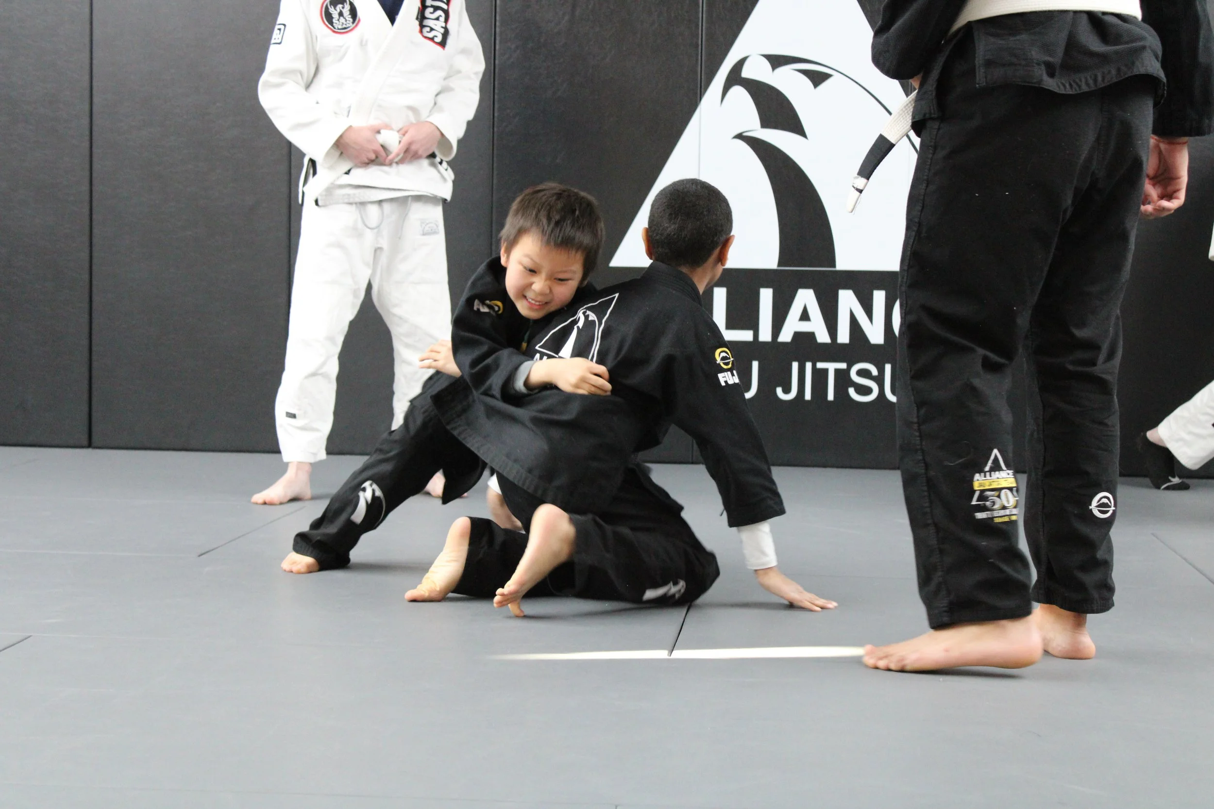 Two children practicing Brazilian Jiu-Jitsu on a mat during a class, with one child on top of the other gripping his opponent's collar, while a third child stands nearby. All are wearing black or white gis with patches.