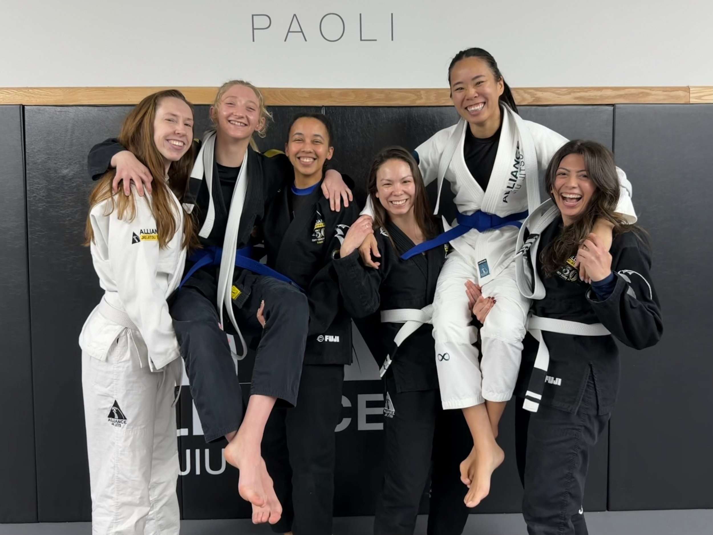 Six women in Brazilian Jiu-Jitsu gis smiling and posing together in a gym. Some are wearing black belts, and one is being lifted by another.
