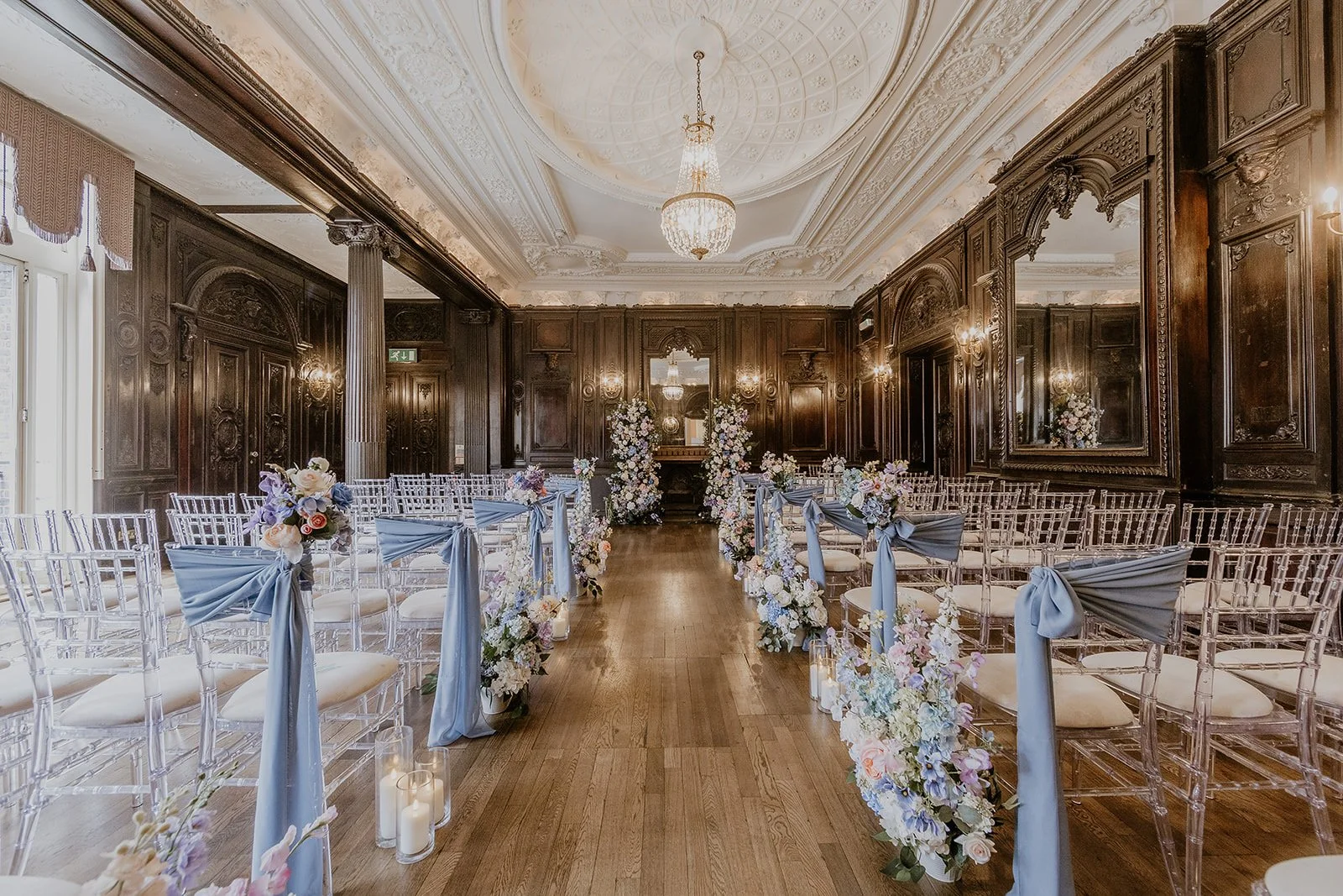 An elegant wedding ceremony setup in a grand, ornate room with dark wood paneling, chandeliers, and floral decorations. Clear chairs with light blue sashes line both sides of the aisle, with flower arrangements and candles along the walkway.