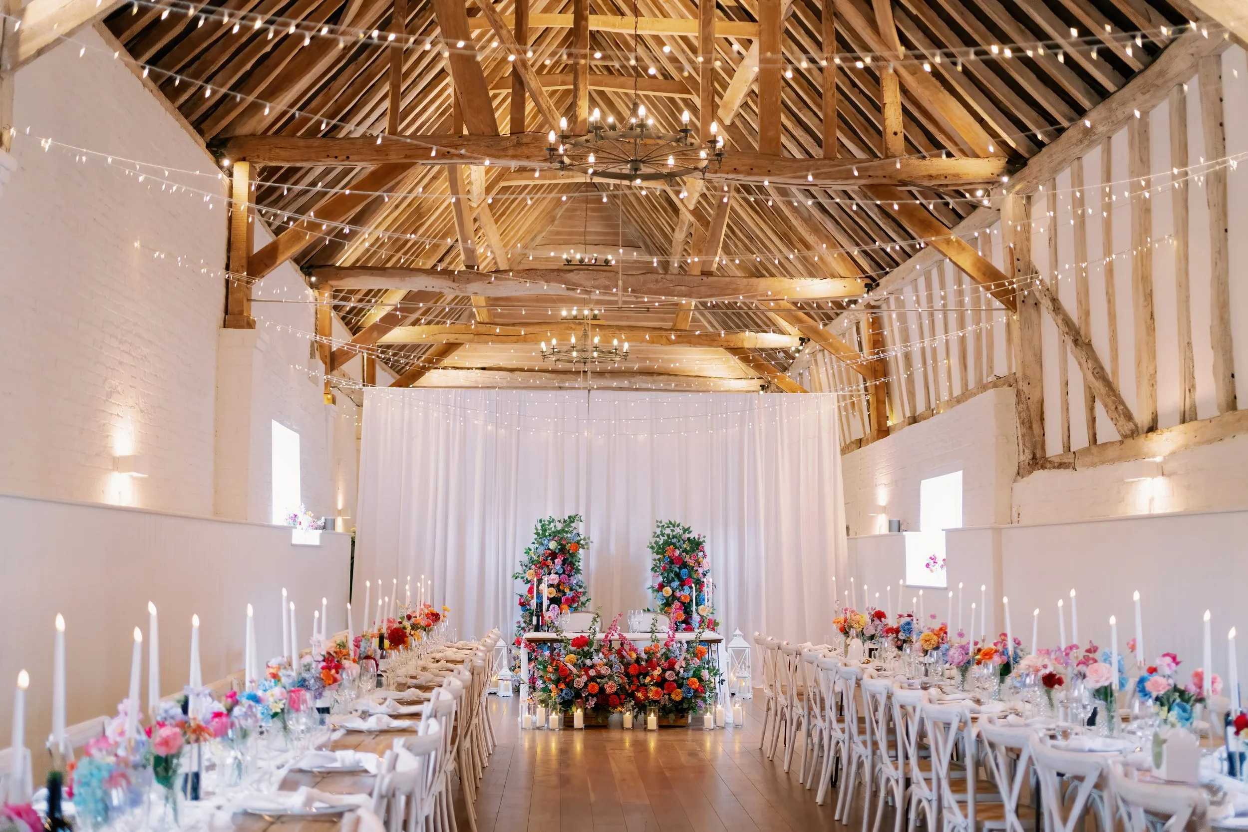 Wedding reception decorated with floral arrangements, candles, and fairy lights in a rustic barn with wooden beams and white walls.