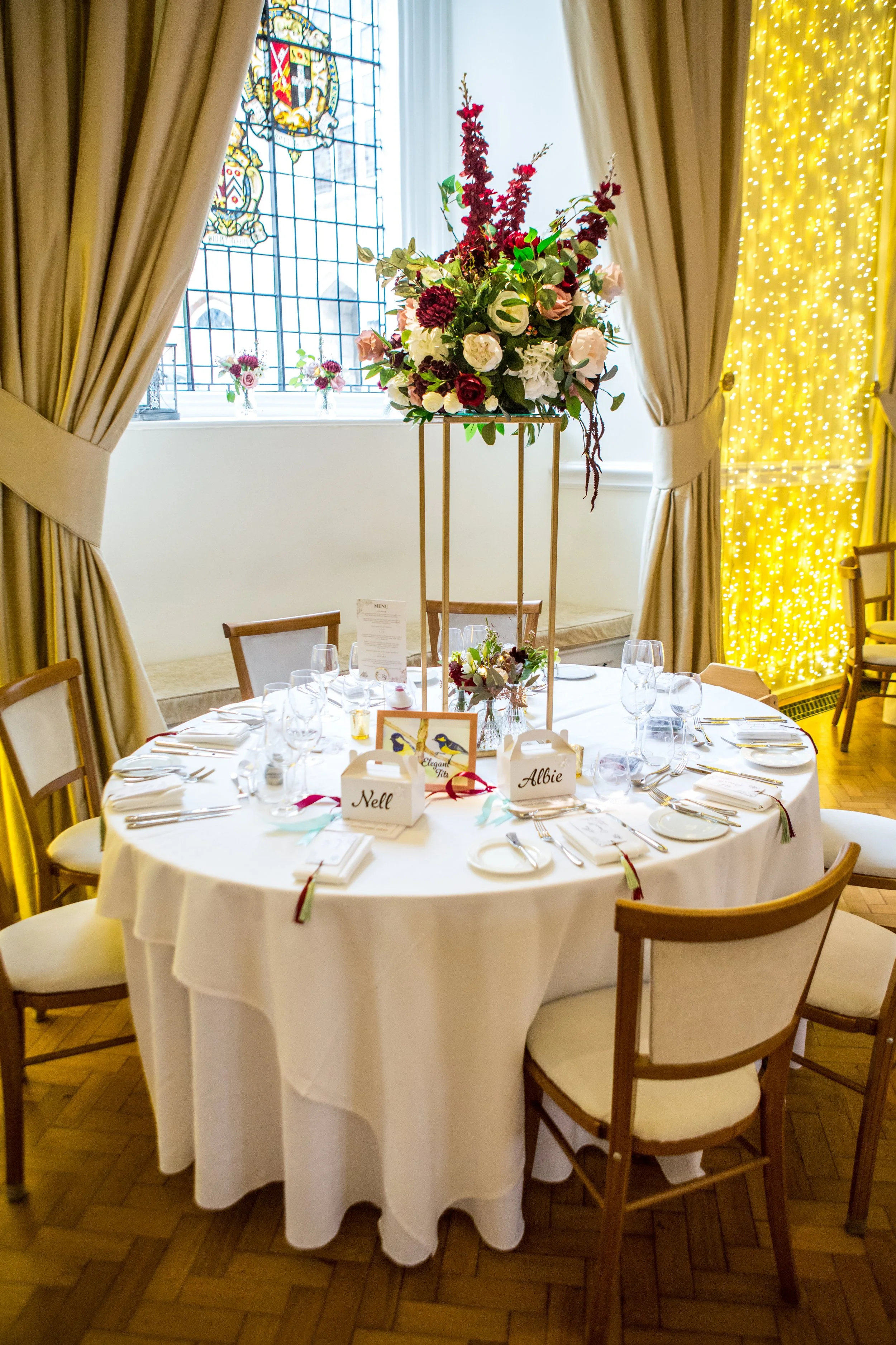 Round dining table with white tablecloth, decorated with a tall floral centerpiece, set with glassware, plates, and silverware, surrounded by six chairs, in a bright room with stained glass window and yellow fairy lights.