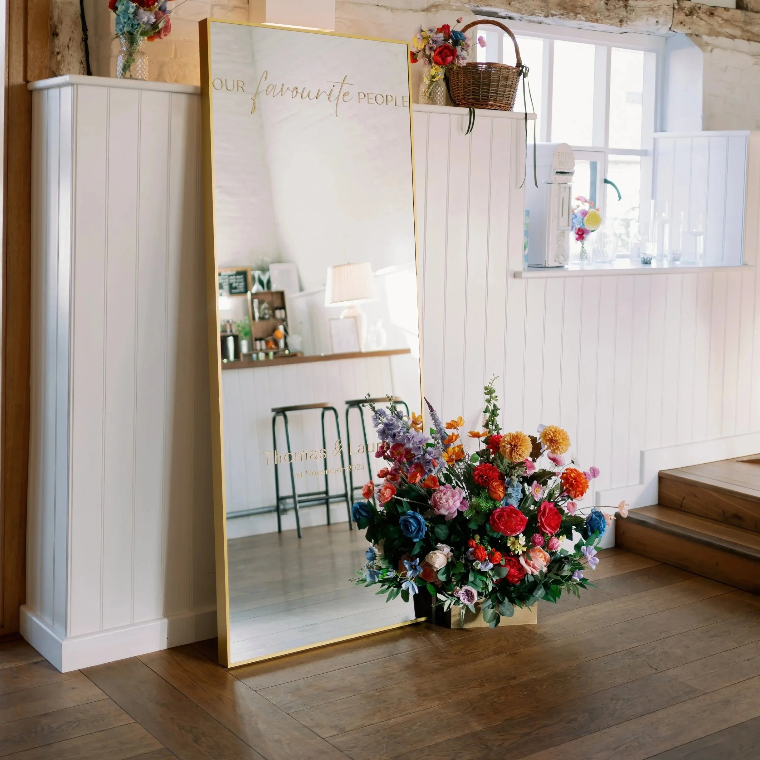 A large mirror leaning against a white paneled wall next to a floral arrangement and a small step leading up to a counter in a bright room.