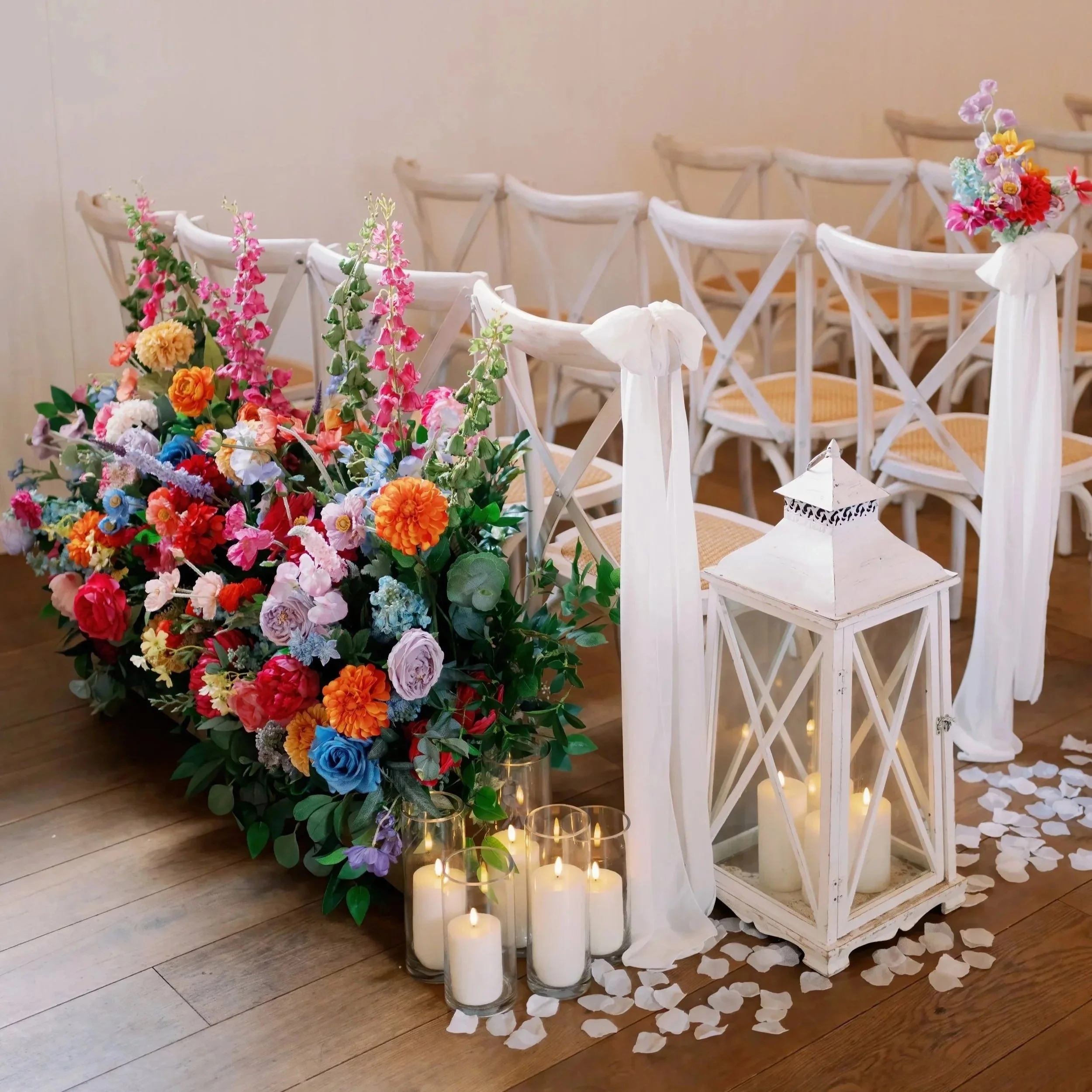 White chairs with white fabric decorations, colorful flower arrangements, white lantern, candles, and flower petals on the floor, set up for a wedding ceremony.