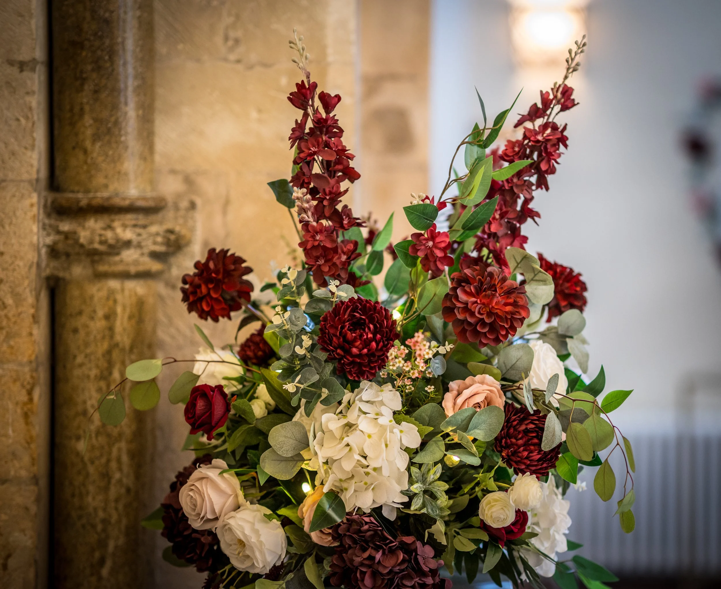 A colorful floral arrangement with red, white, and pink flowers, green leaves, and small white accents, positioned indoors near a stone wall.