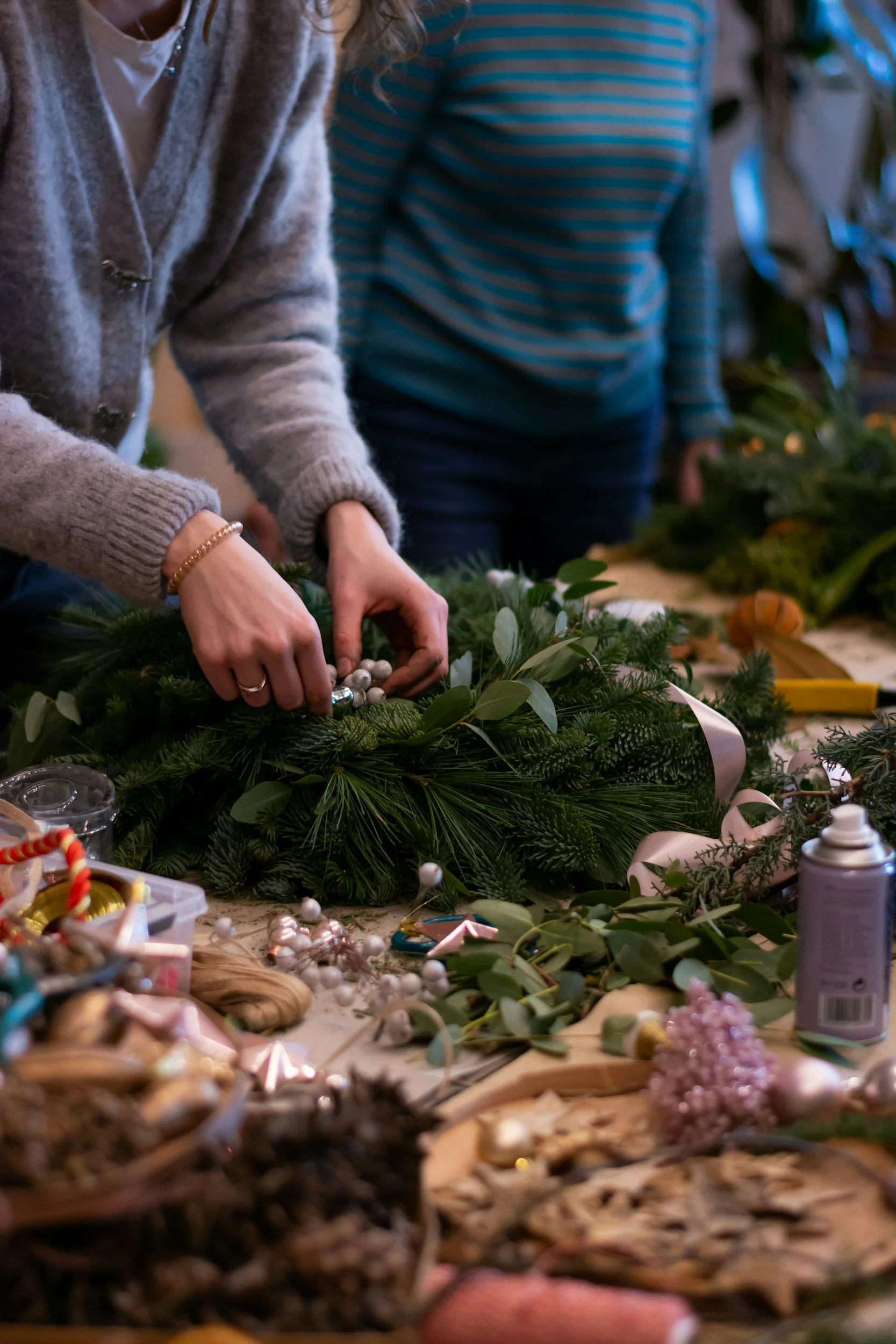 People assembling a holiday wreath using greenery, pinecones, and decorations on a wooden table, with crafting supplies scattered around.