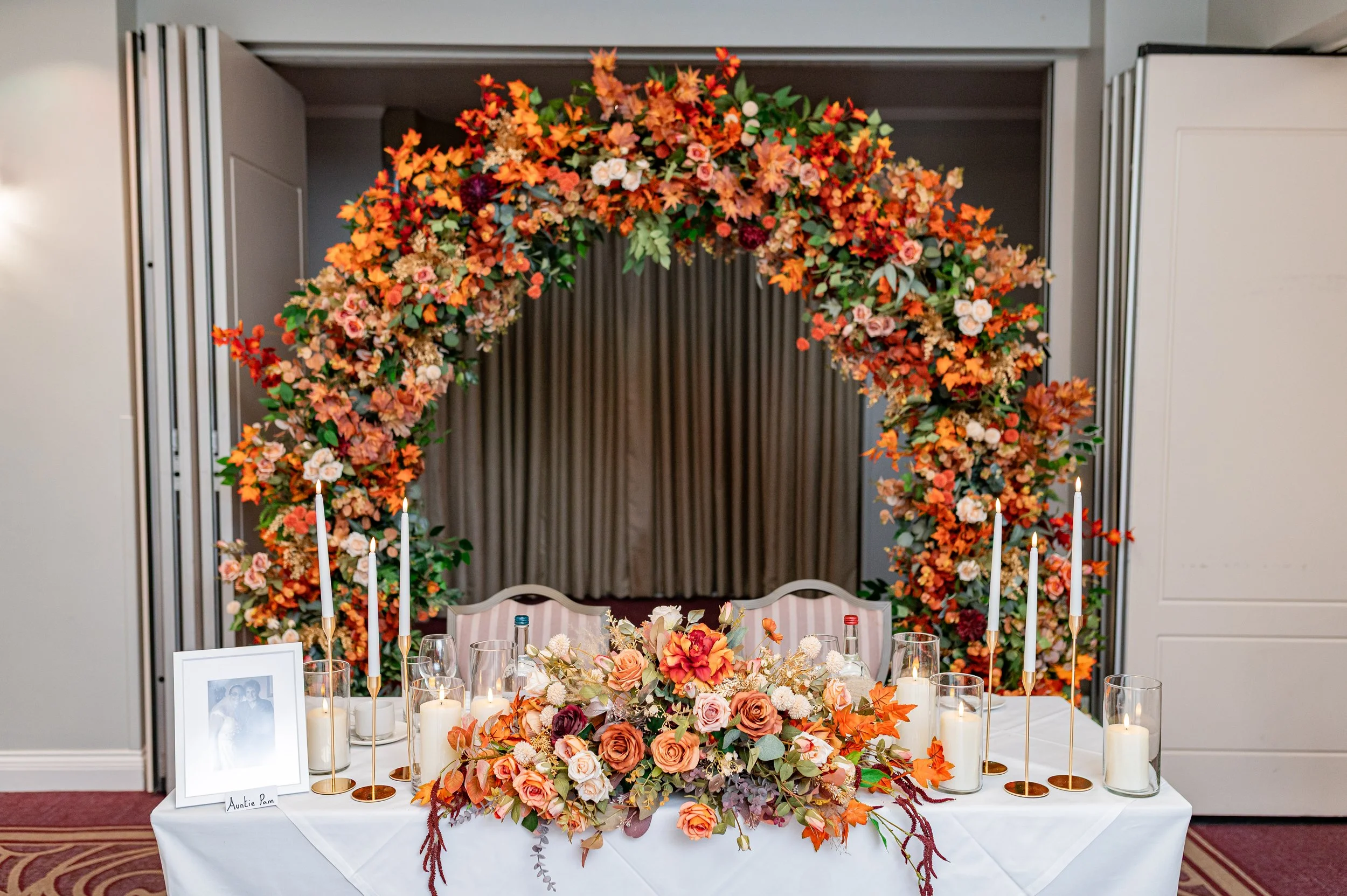 A decorated wedding reception table with a floral centerpiece, candles, and a framed photograph, against a backdrop of a large floral arch with orange, pink, and red flowers.