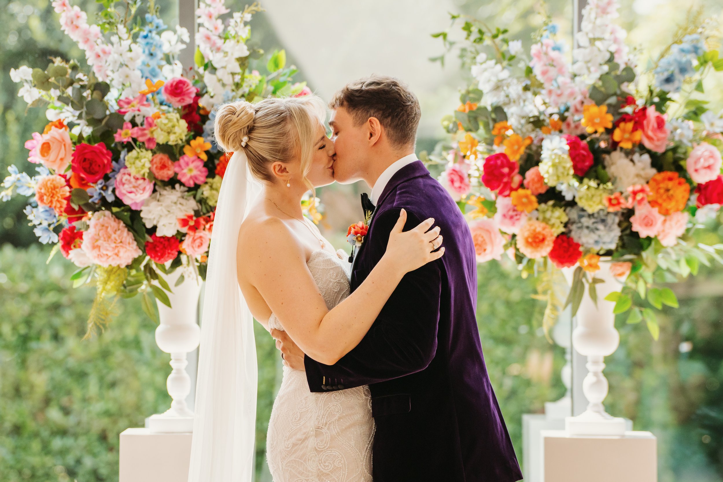 A bride and groom sharing a kiss at their wedding altar with colorful floral arrangements in the background.