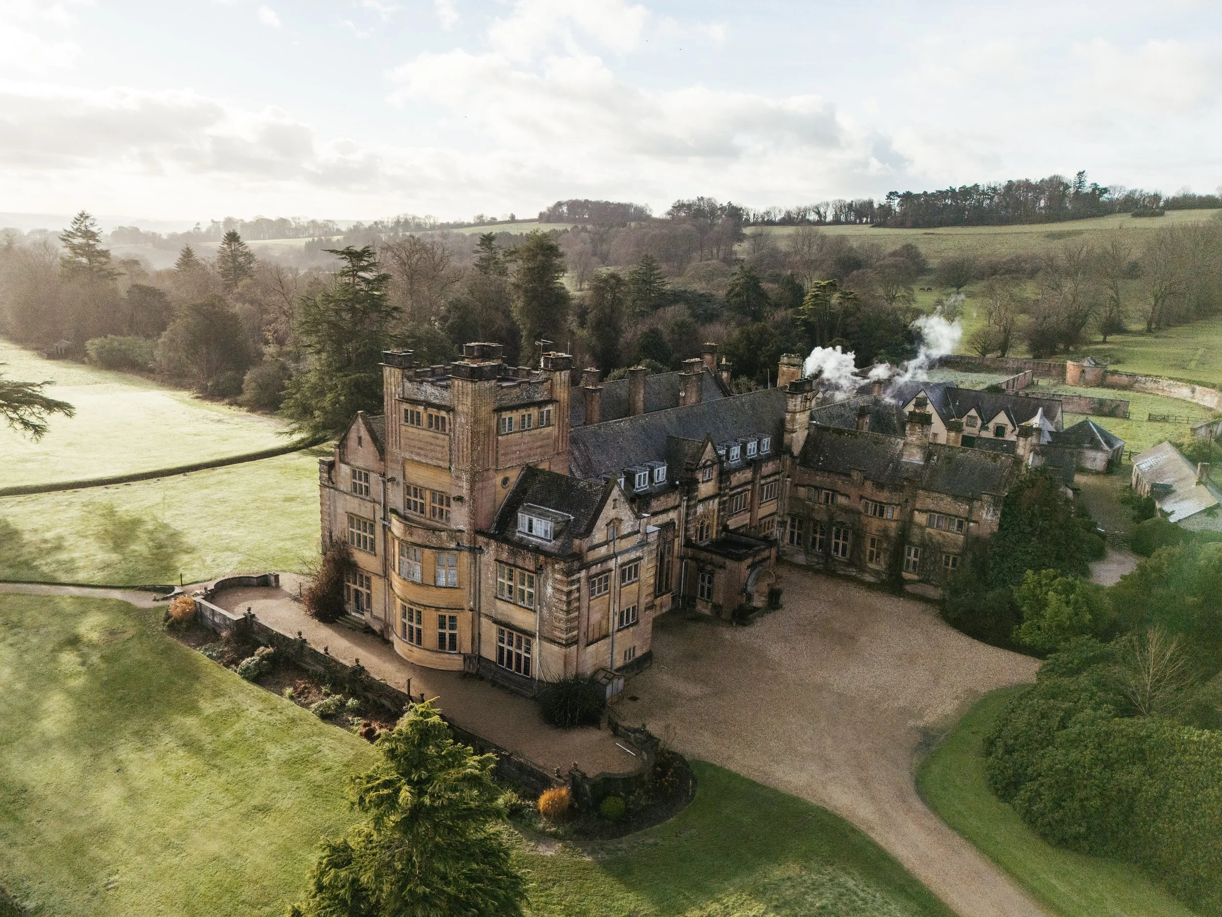 Aerial view of an old stone mansion surrounded by expansive green lawns and trees, with a few buildings and smoke rising from the chimneys in the background.