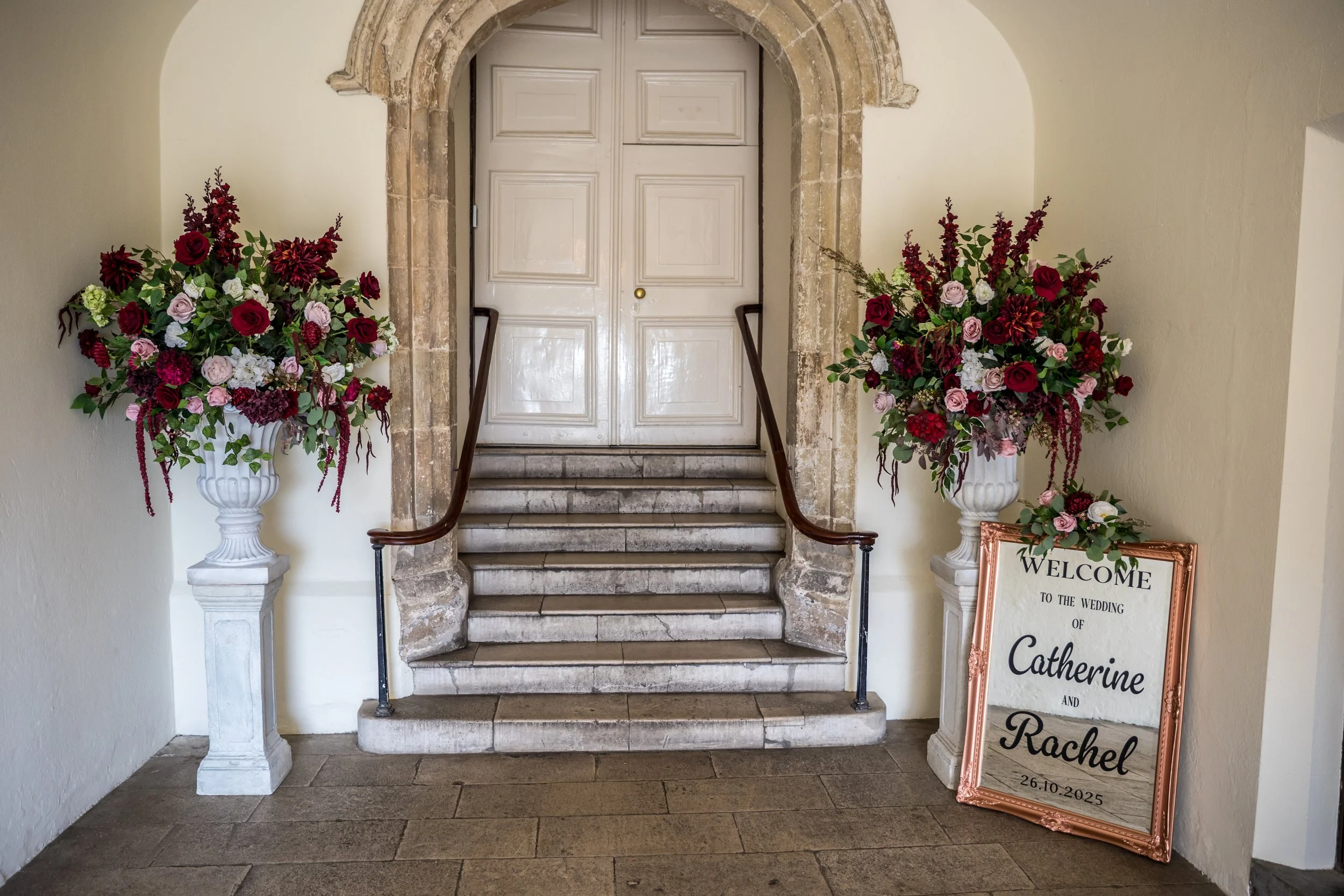 Decorative wedding entrance with two large floral arrangements in white urns on pedestals, a welcome sign for the wedding of Catherine and Rachel with the date 26.10.2025, and a staircase leading to a closed door.