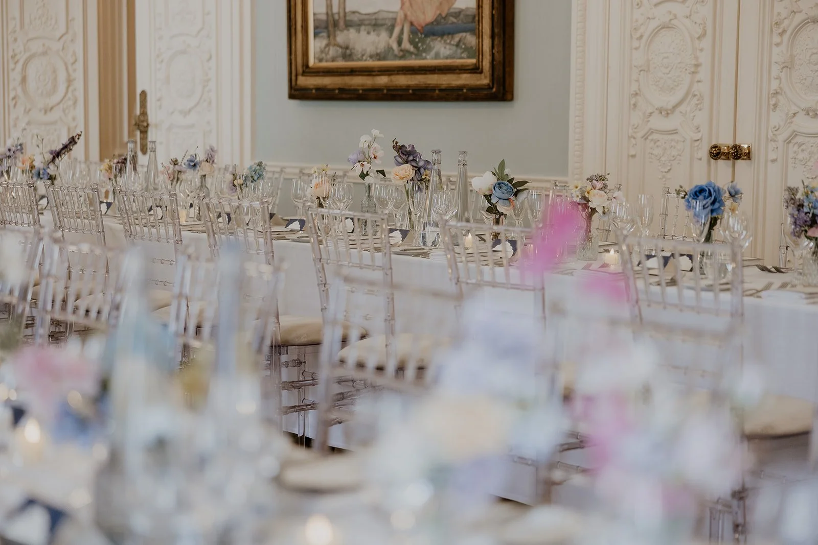 Elegant banquet table set with floral centerpieces, clear glassware, and gold-colored chairs in a decorated room with ornate wall detailing and a framed painting.