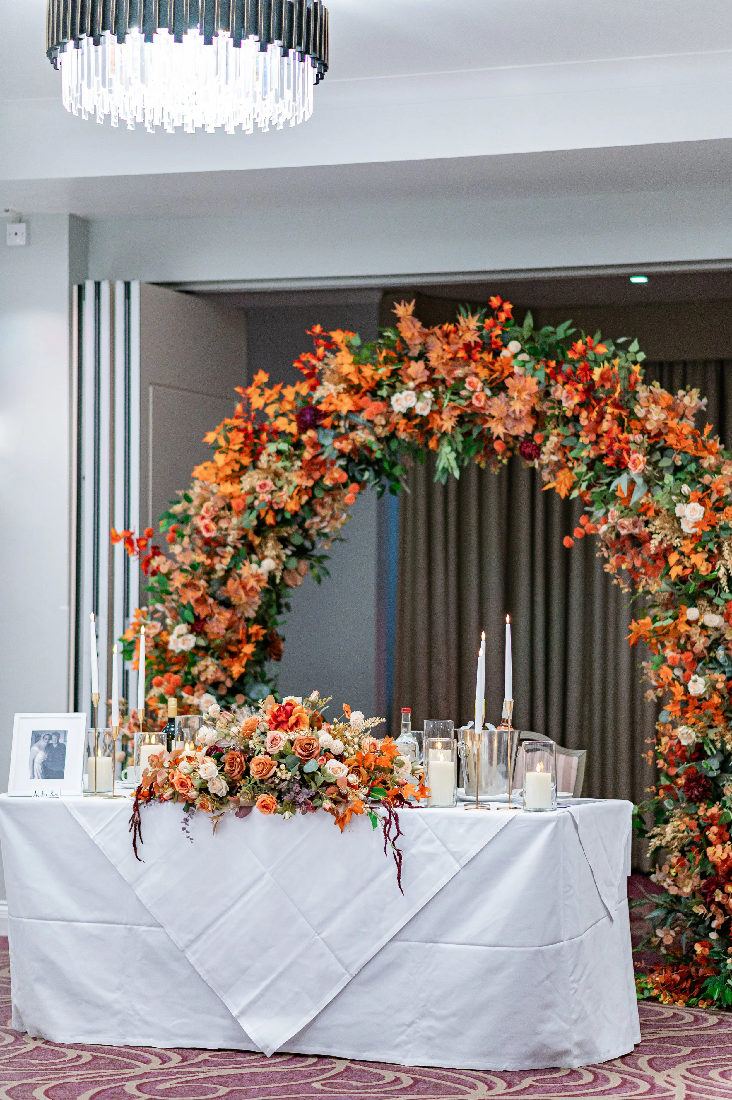 A decorated table with a floral arrangement, candles, and a black-and-white framed photo in front of a large fall-themed floral arch, at a formal event or wedding reception.