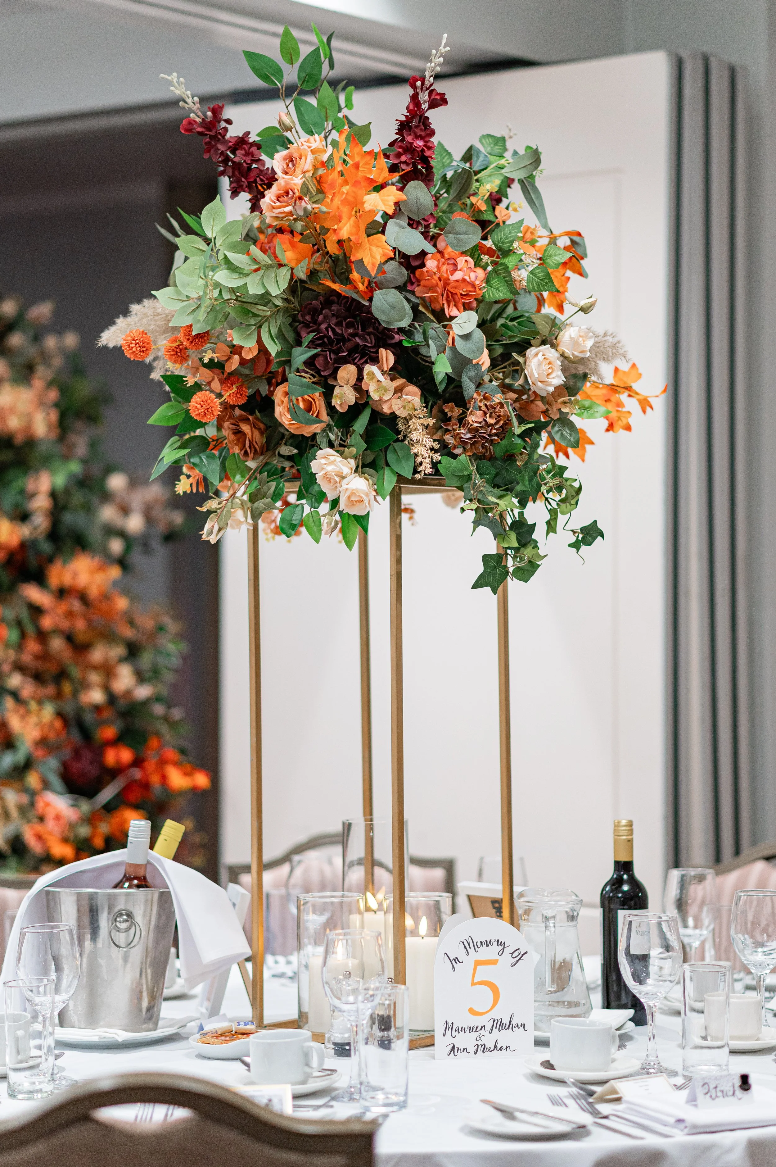 A tall floral centerpiece with orange, peach, deep red, and cream flowers and green foliage, on a banquet table with glassware, plates, and a table number sign. The sign indicates a memory of Maureen Muhan and Ann Muhan, with the number 5.