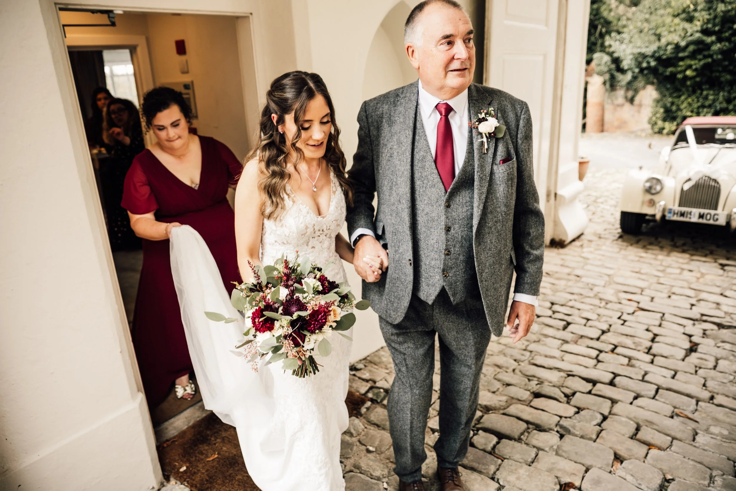 A bride in a white wedding dress holding a bouquet of flowers, walking outdoors with a man in a gray suit, surrounded by a cobblestone pathway and vintage cars in the background.