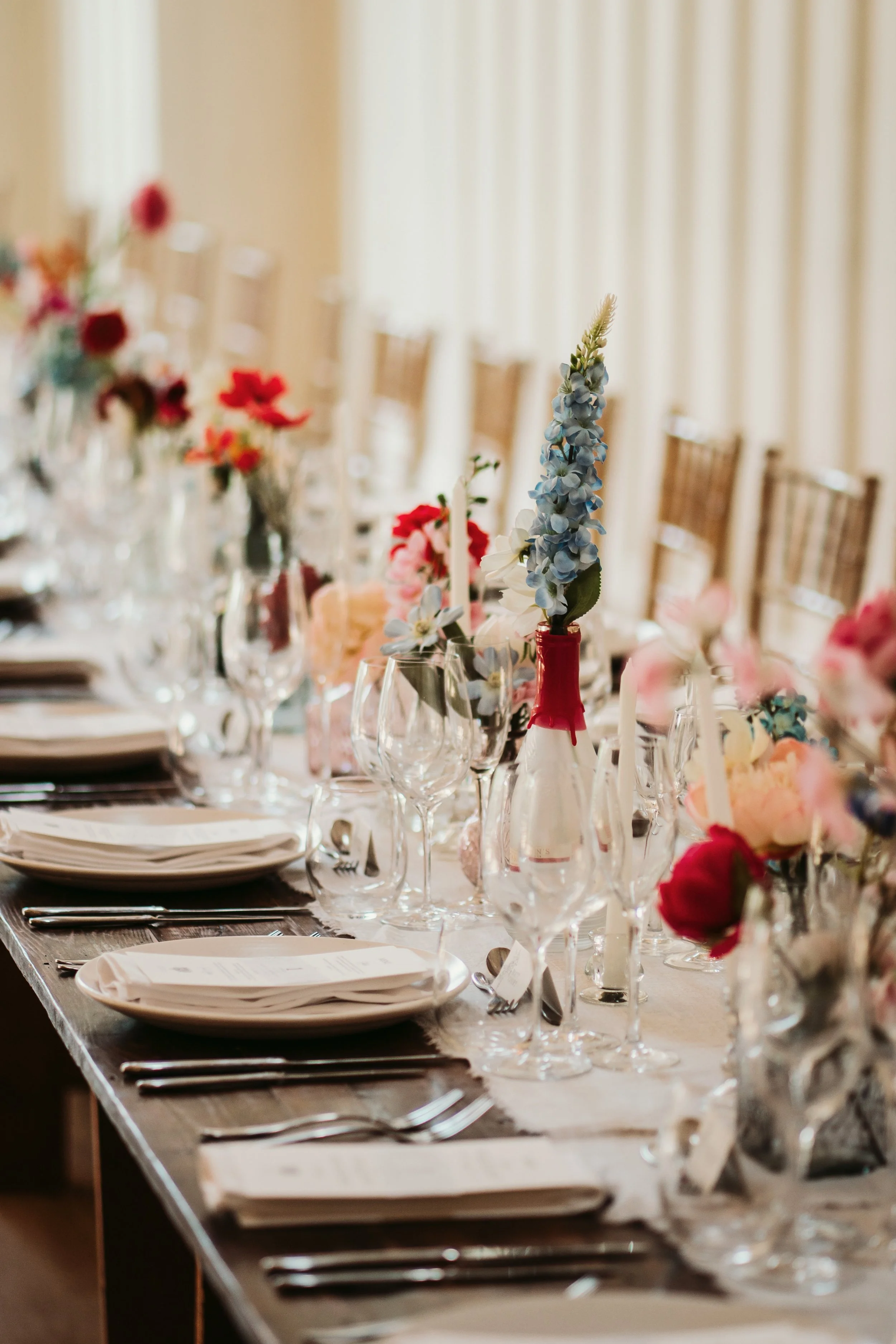 A long banquet table decorated with elegant glassware, plates, silverware, and colorful floral arrangements in vases, set for a formal event.