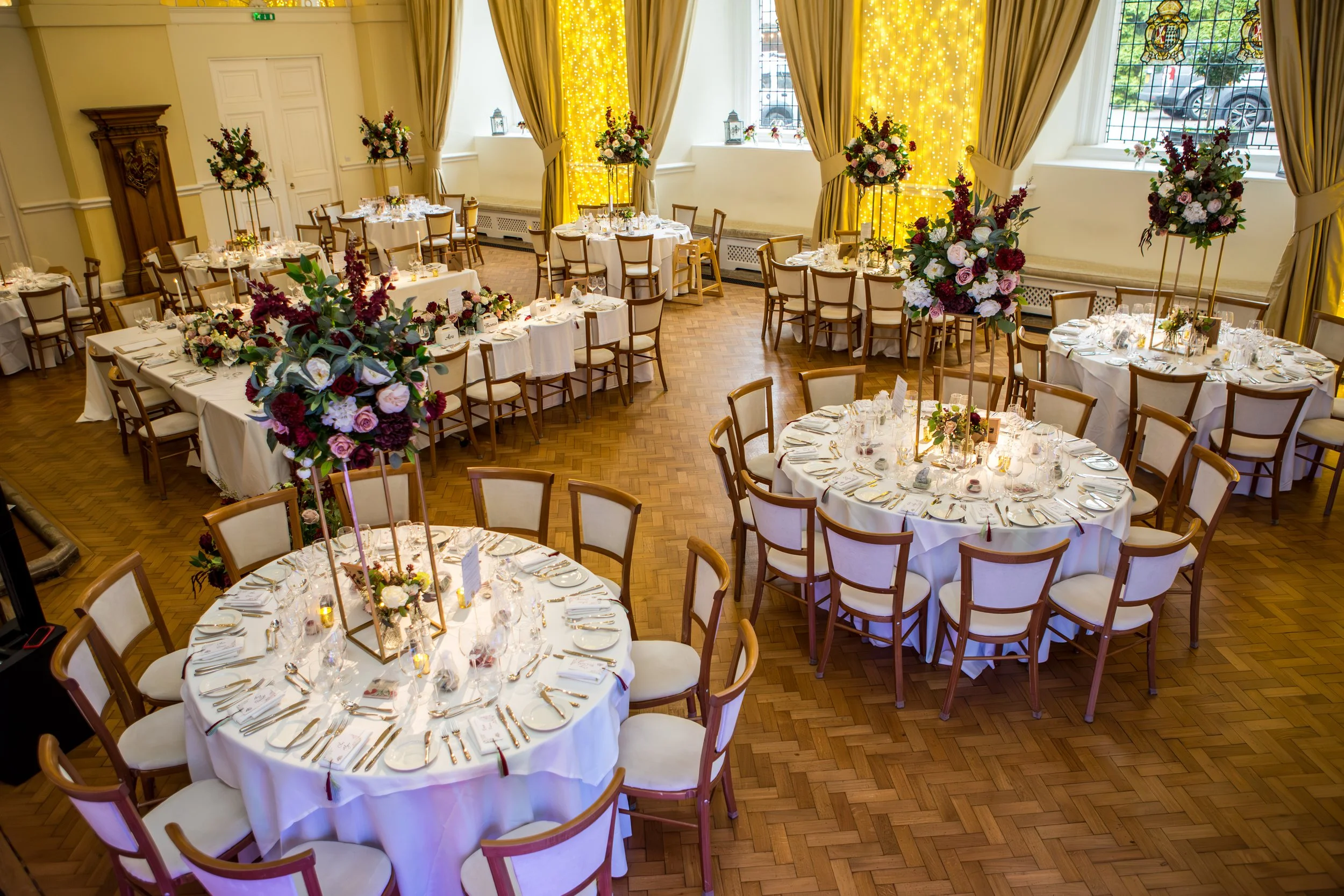 Elegant banquet hall decorated with round and rectangular tables covered in white tablecloths, set with fine dinnerware and glassware, and adorned with tall floral centerpieces featuring roses and other flowers in shades of pink, purple, and white. T