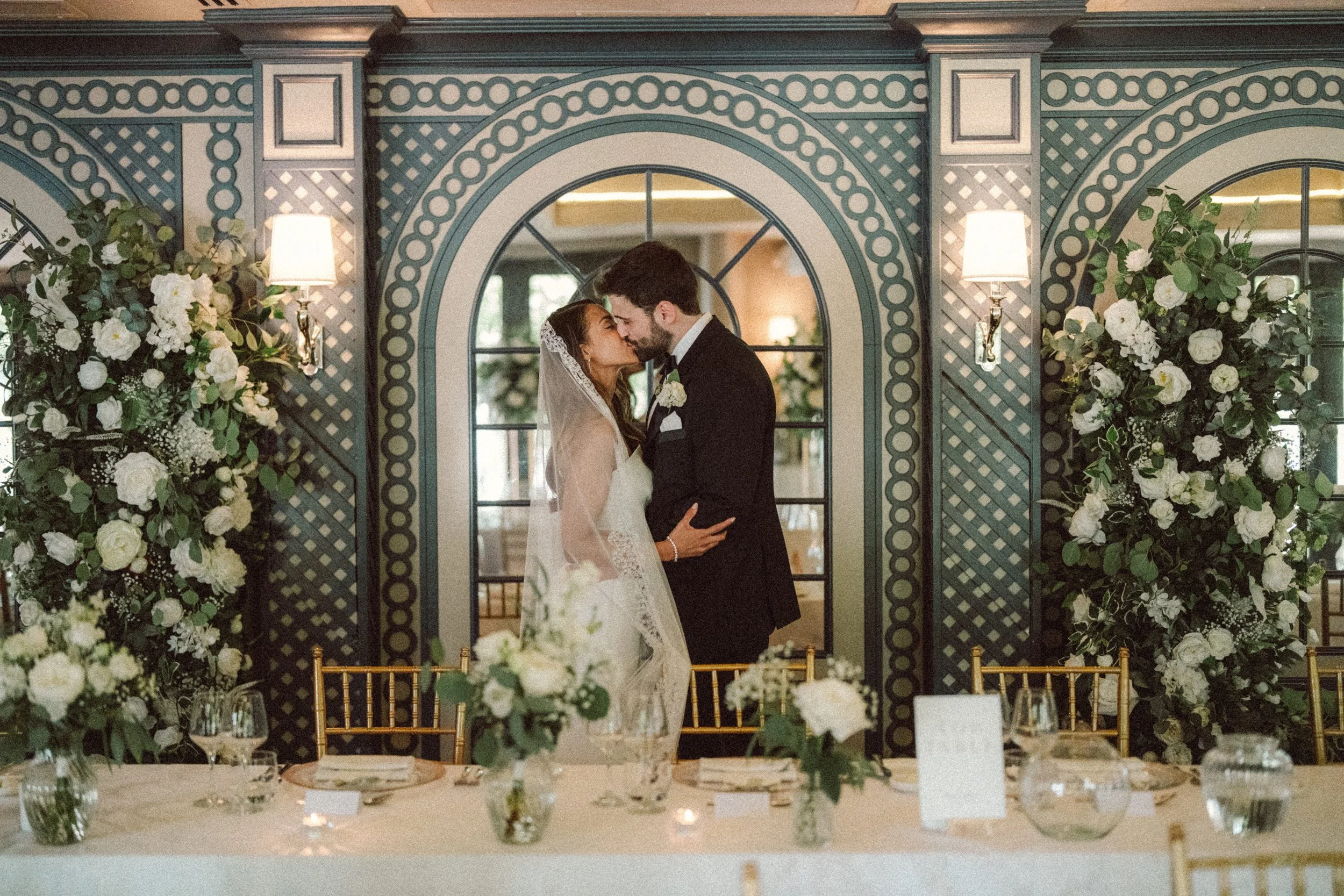 A bride and groom sharing a kiss at their wedding reception, standing behind a decorated table, with floral arrangements and a patterned wall in the background.