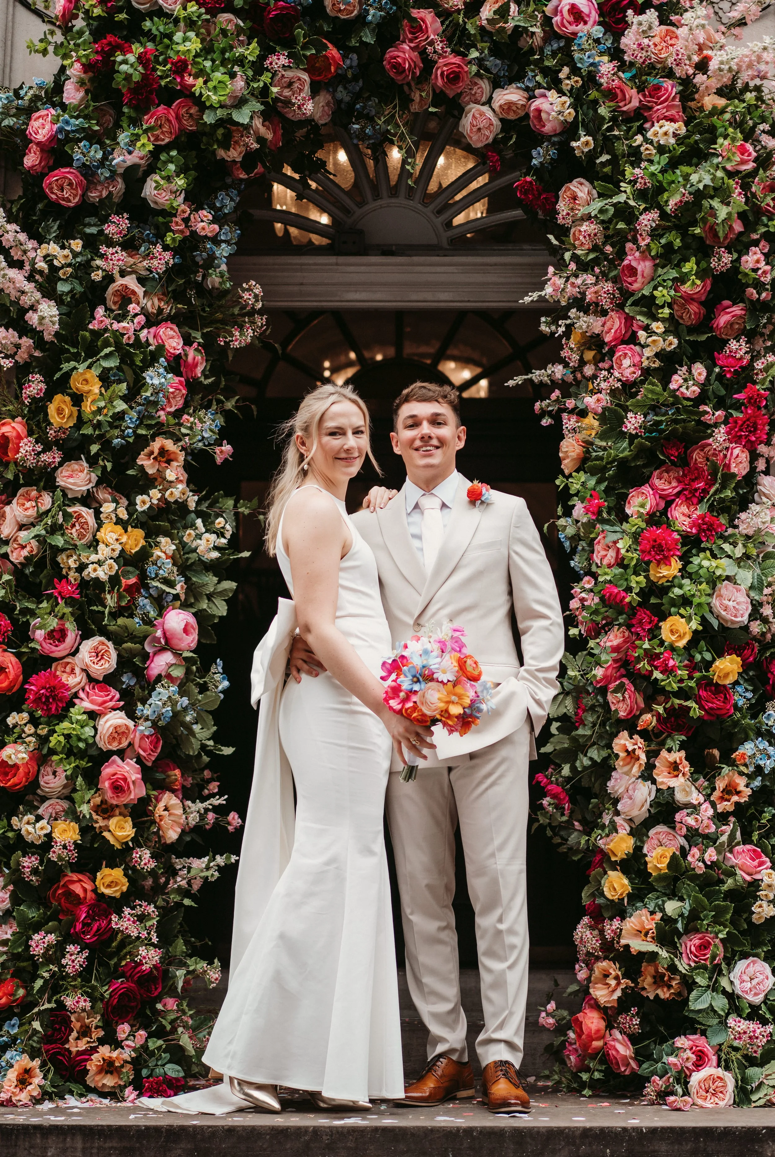 A newlywed couple standing under a floral arch, smiling, with the bride holding a colorful bouquet, dressed in white, and the groom in a light-colored suit, indoors with warm lighting.