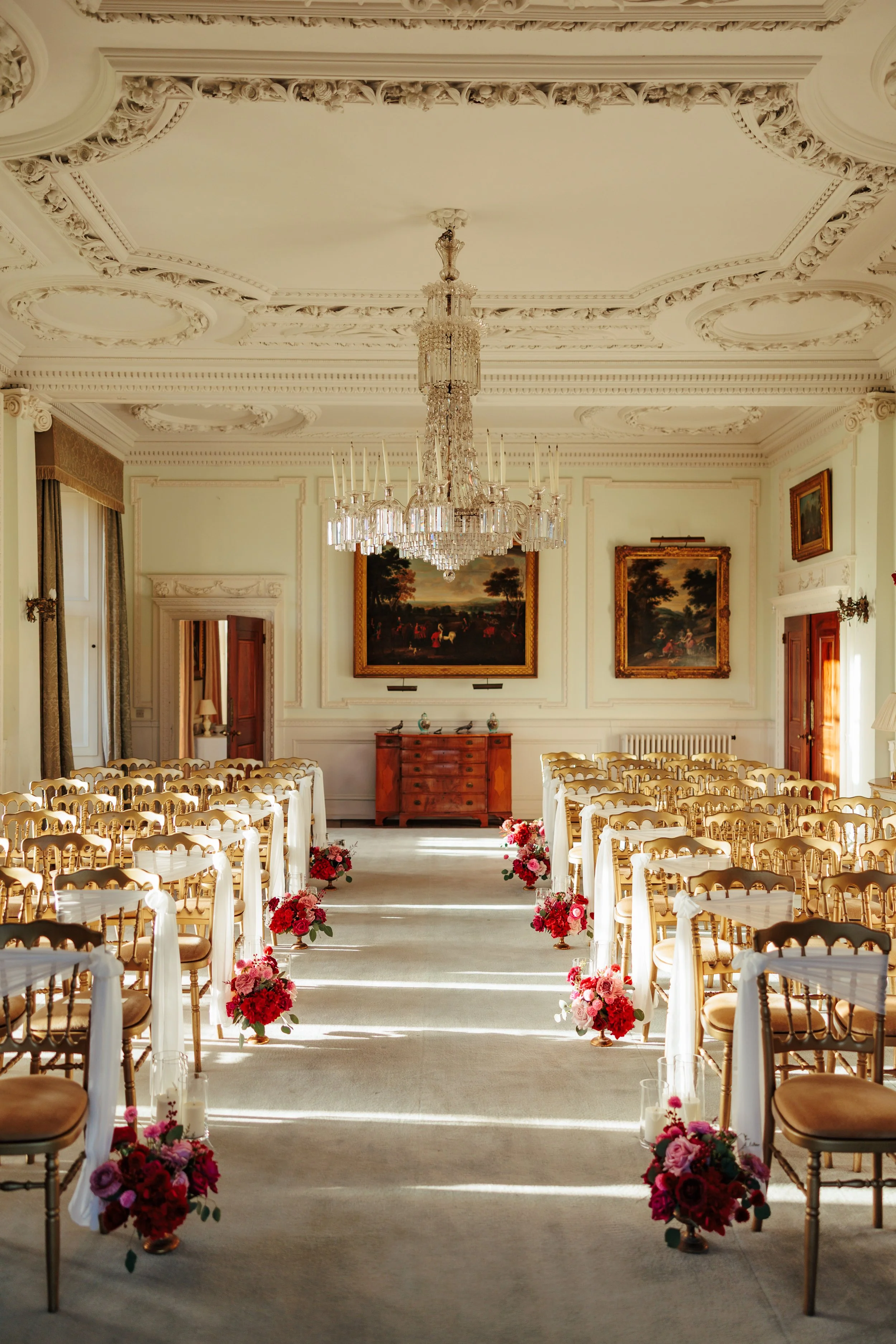 Elegant wedding venue with rows of gold chairs decorated with pink and red flower arrangements, a chandelier hanging from a decorated ceiling, and paintings on the walls.