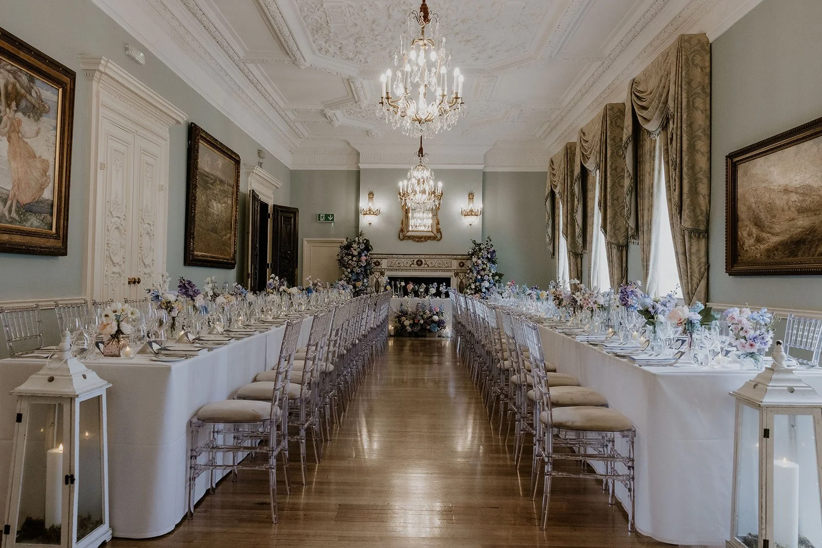 Elegant wedding reception hall with long tables decorated with flowers and glassware, chandeliers hanging from an ornate ceiling, and large windows with draped curtains.
