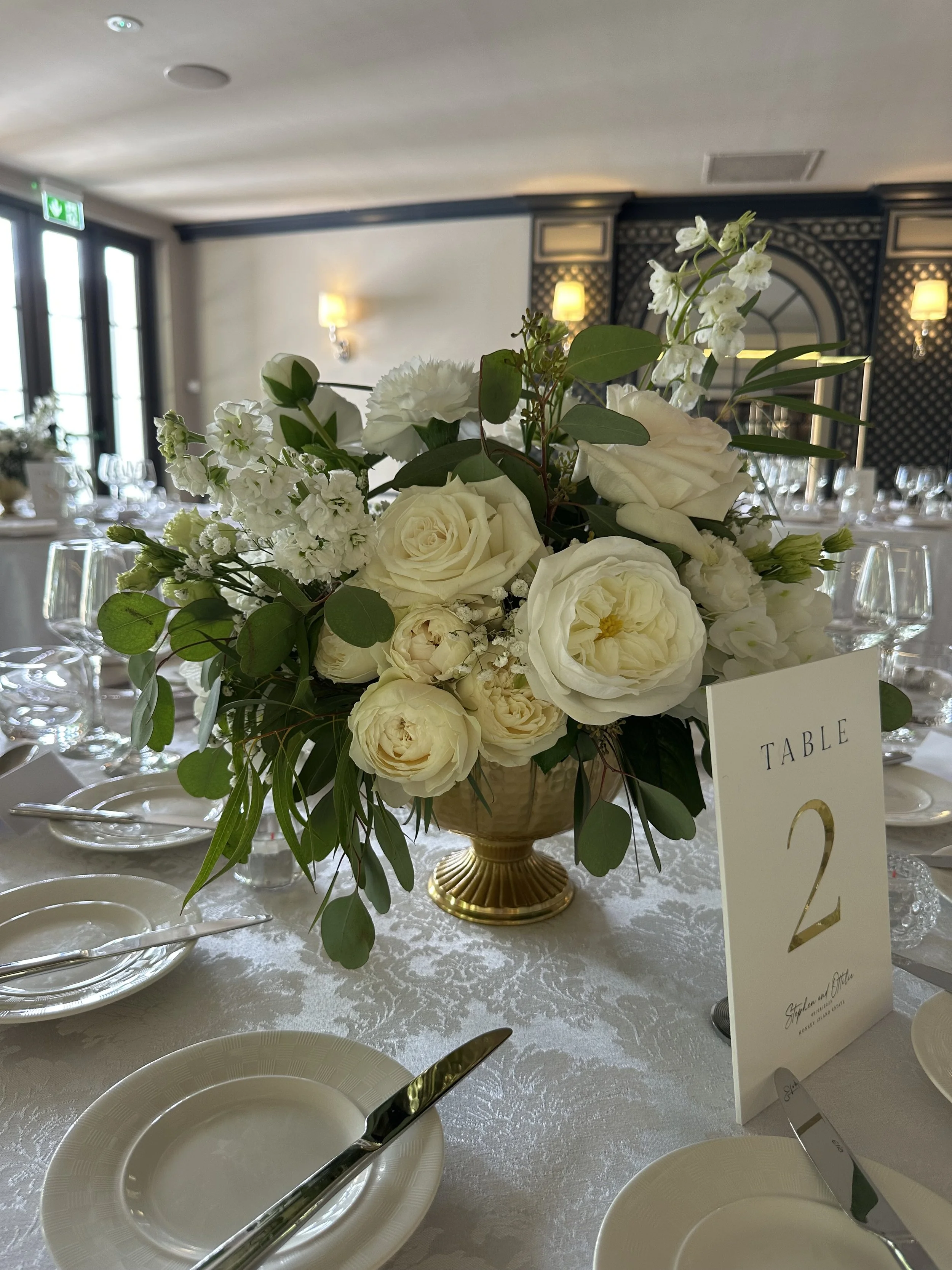 Elegant floral centerpiece with white roses and greenery on a formal dining table, with table number card reading '2'.
