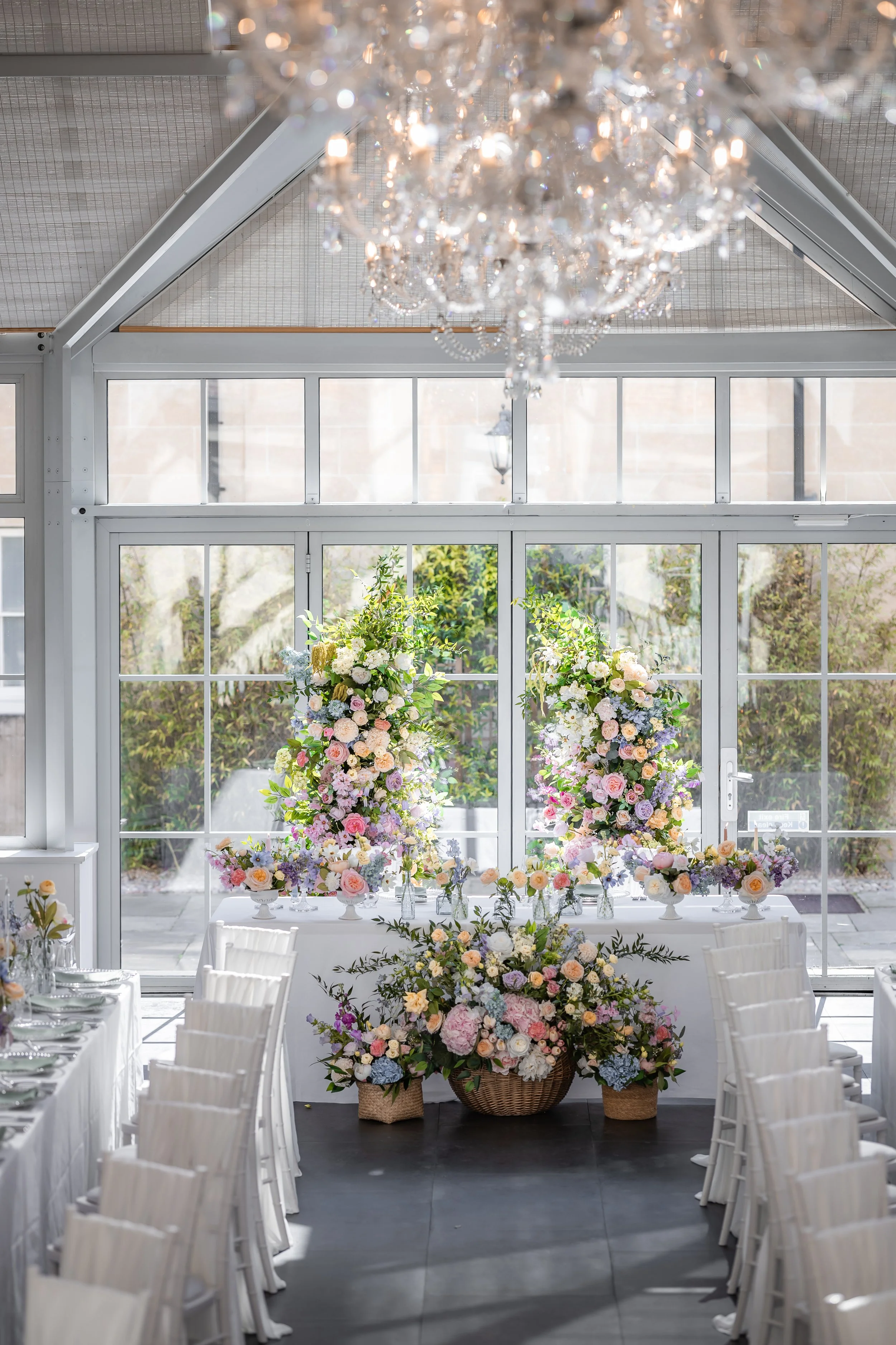 Elegant indoor event space decorated with large floral arrangements, white chairs, and a crystal chandelier, with glass doors and windows revealing greenery outside.