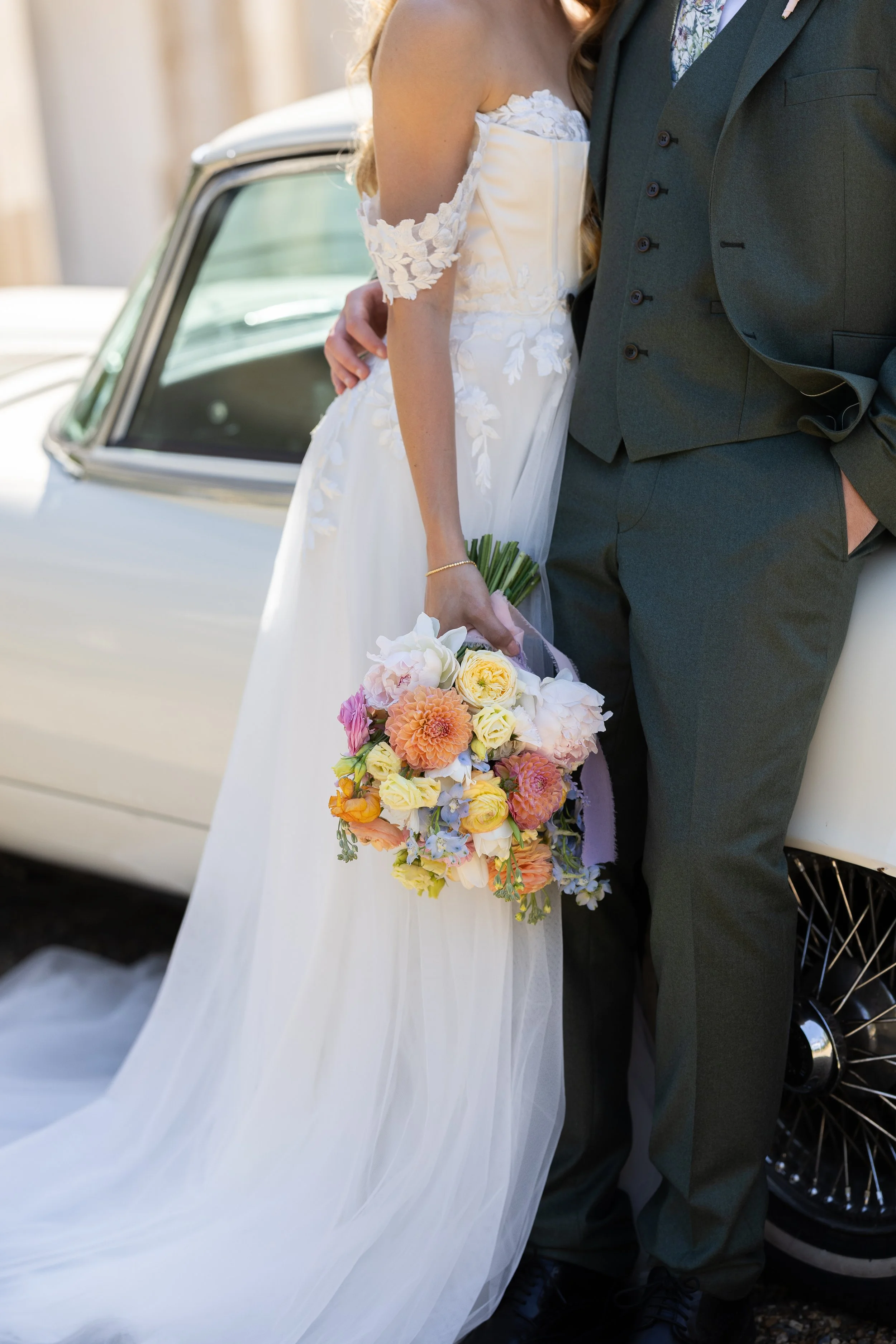 Bride in white wedding dress holding a colorful bouquet leaning against a groom in a dark suit near a vintage white car.