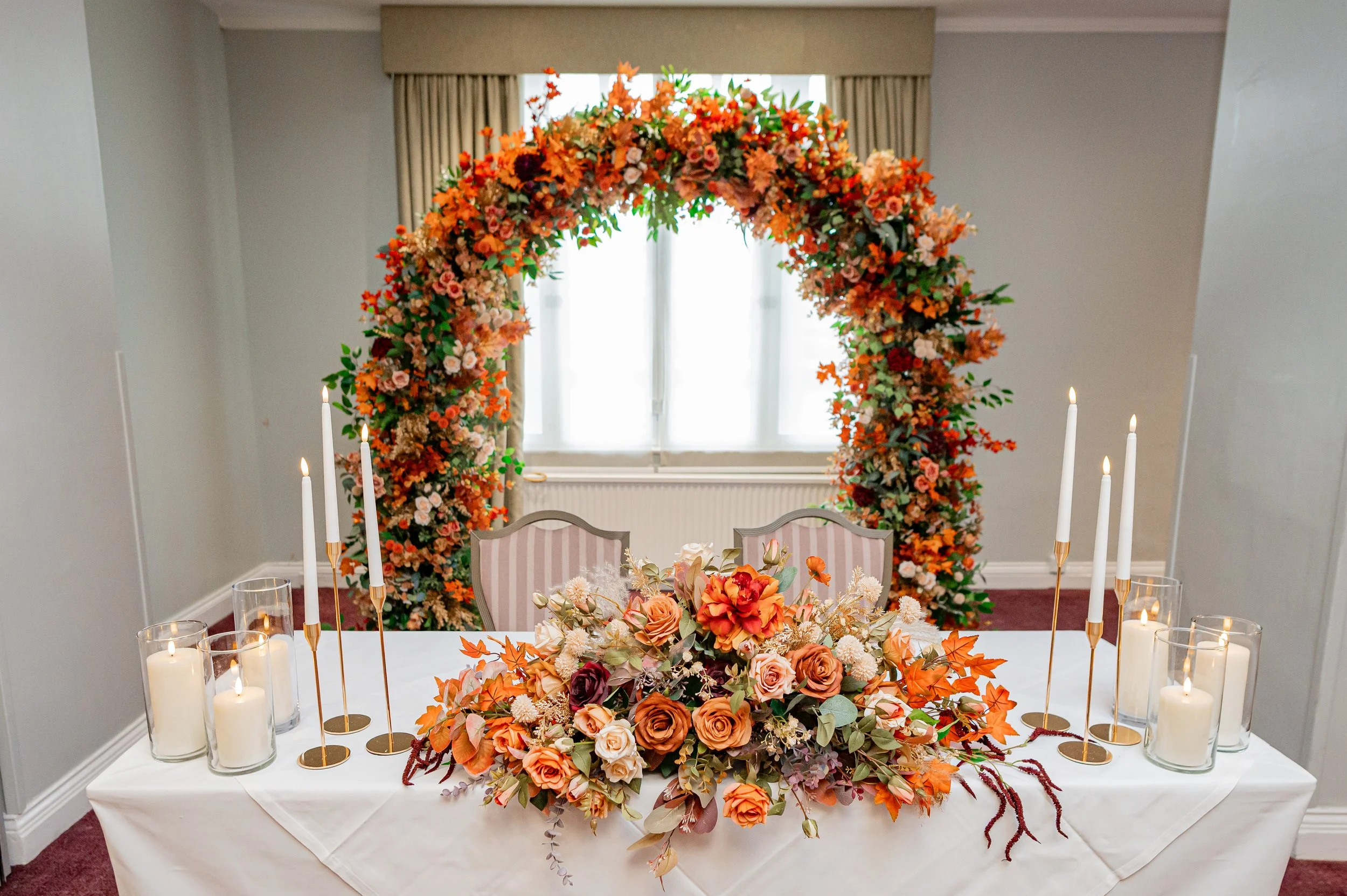 A decorated wedding table with floral arrangements, candles, and an orange flower arch in the background.