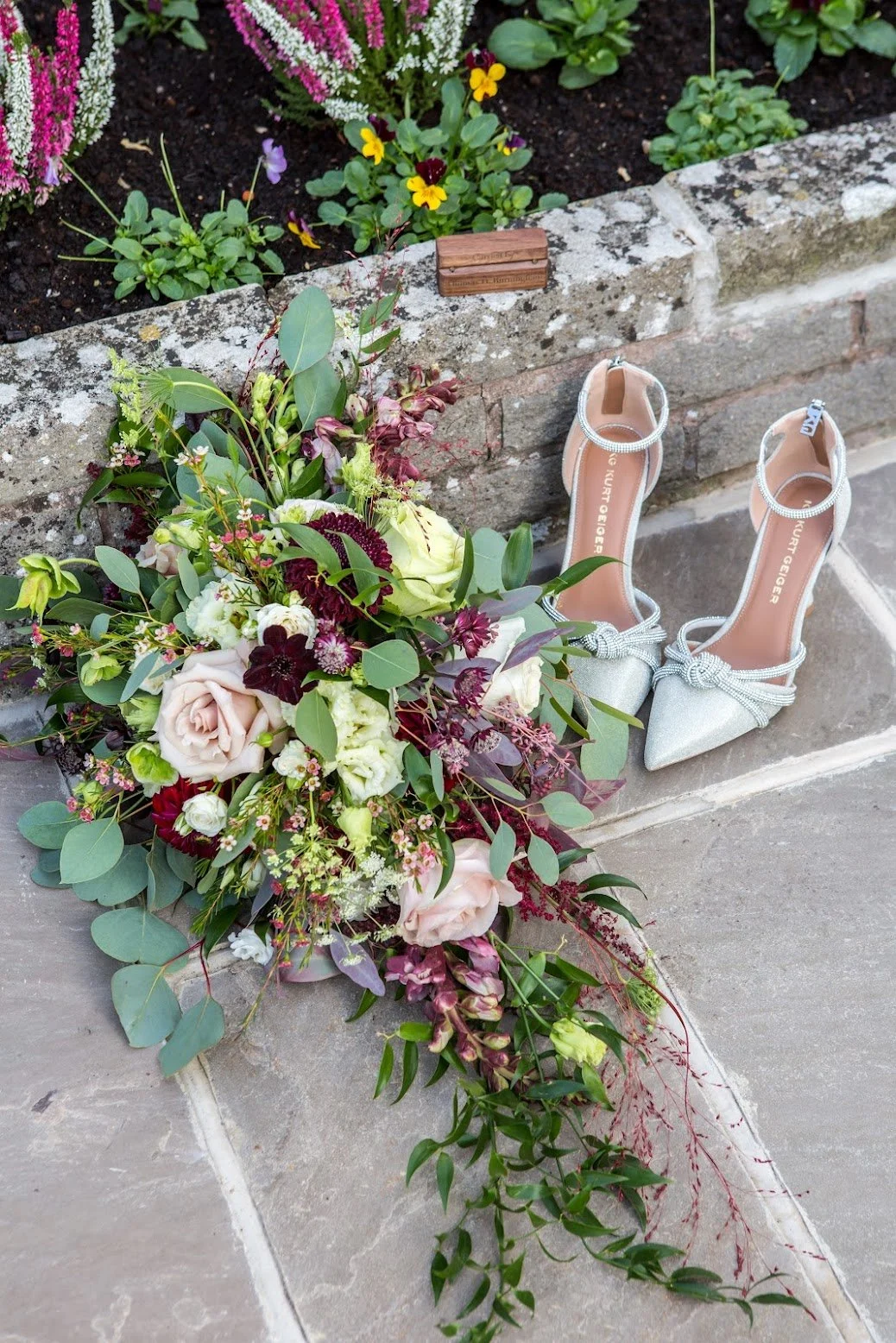 A bouquet of mixed flowers including roses, greenery, and other blooms, resting on a sidewalk near a pair of silver high-heeled shoes with ankle straps, against a brick wall with a flower bed.