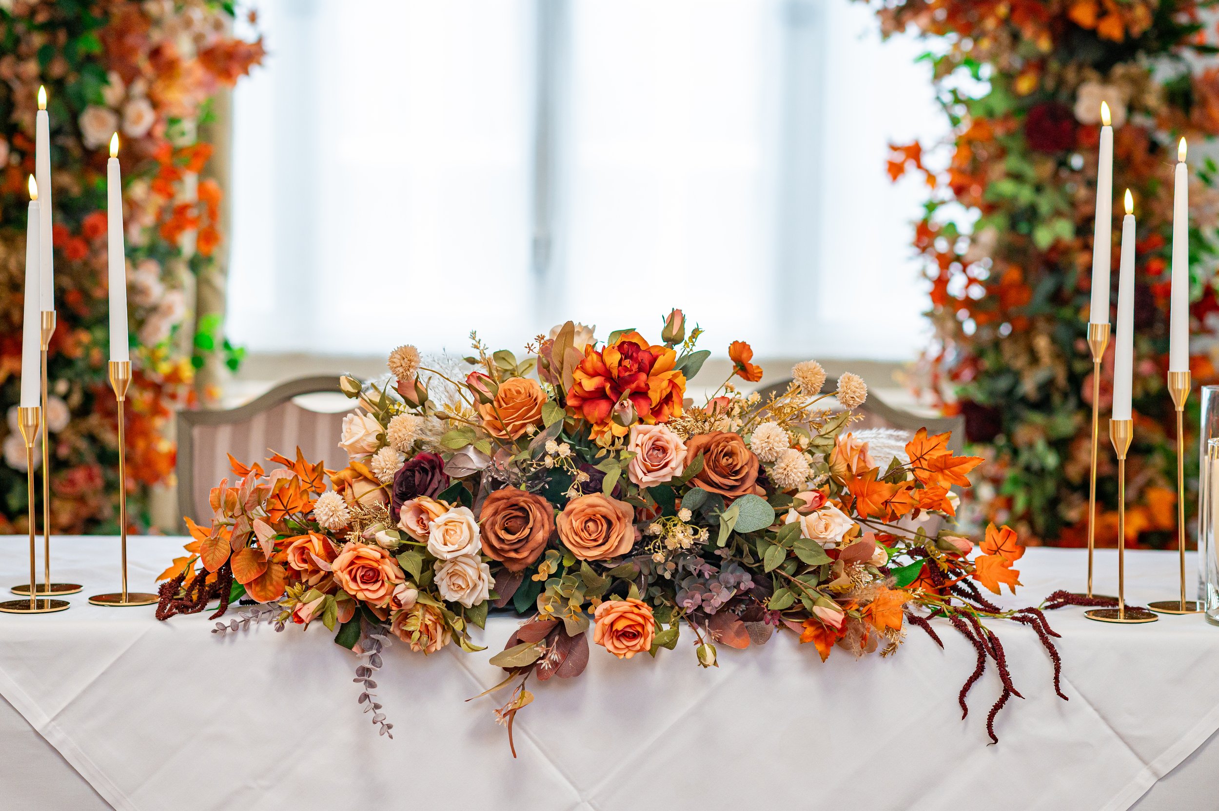 Elegant floral centerpiece with roses and orange flowers on a white tablecloth, surrounded by tall, thin candle holders with lit candles, with a soft background of floral arrangements and a window.