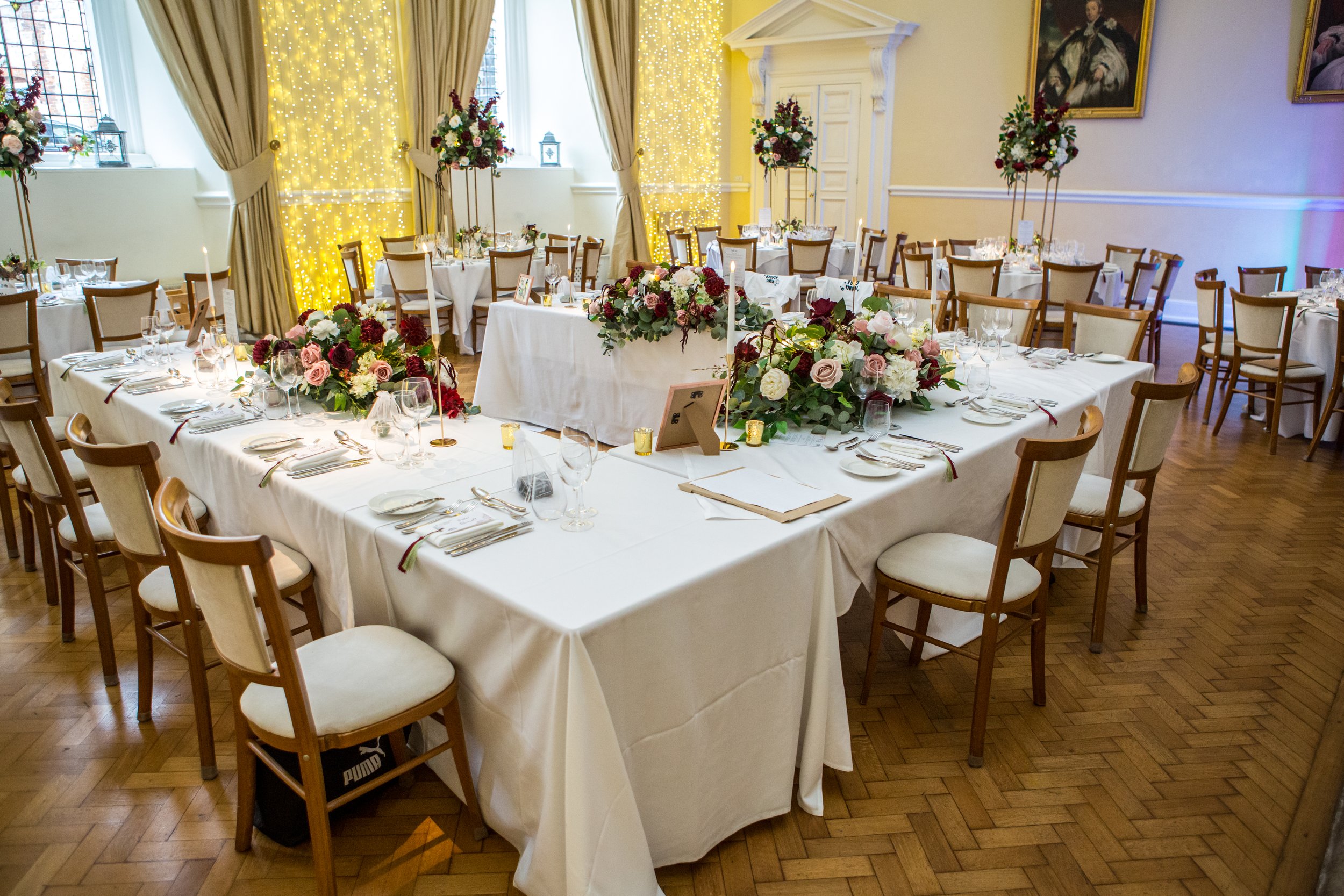 A banquet hall decorated for a wedding reception with multiple tables covered in white tablecloths, adorned with floral centerpieces, candles, and place settings, with string lights and paintings on the walls.
