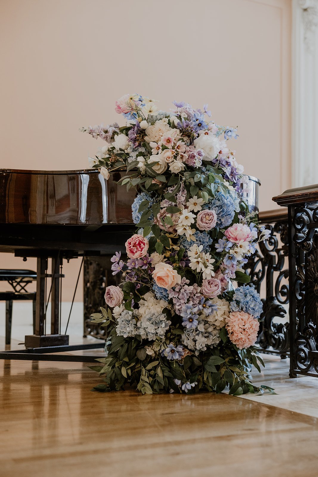 A large arrangement of pink, purple, white, and blue flowers spilling over a black grand piano and a wrought iron railing on a wooden floor.