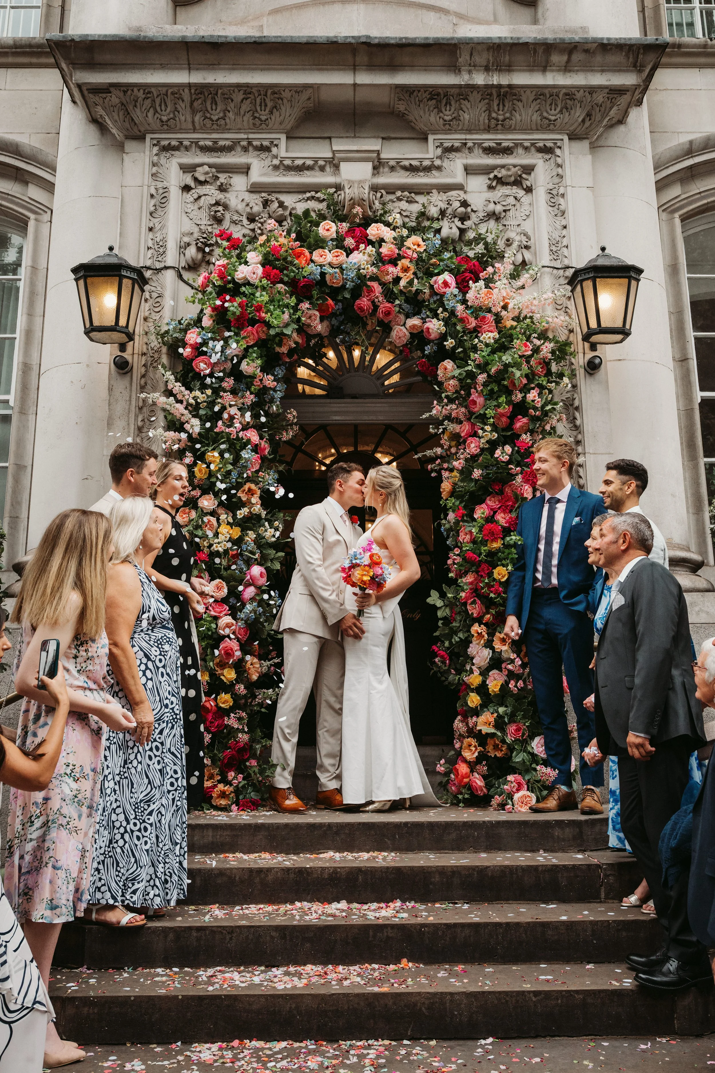A newly married couple shares a kiss on the steps of a building decorated with a large floral arch, surrounded by friends and family celebrating.
