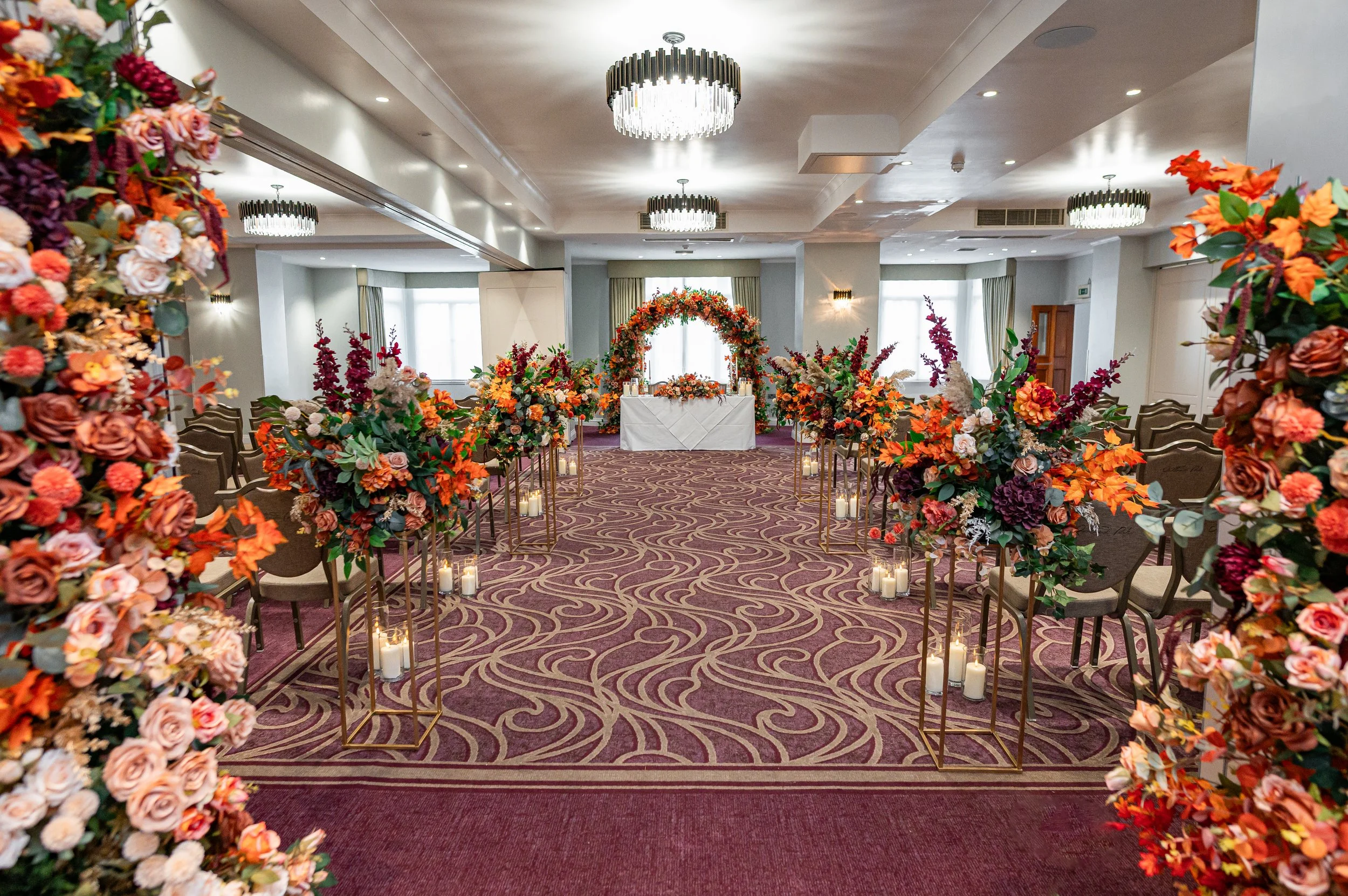 Elegant indoor wedding ceremony setup with floral arches, colorful flower arrangements, candles, and a decorated altar at the front of the aisle.