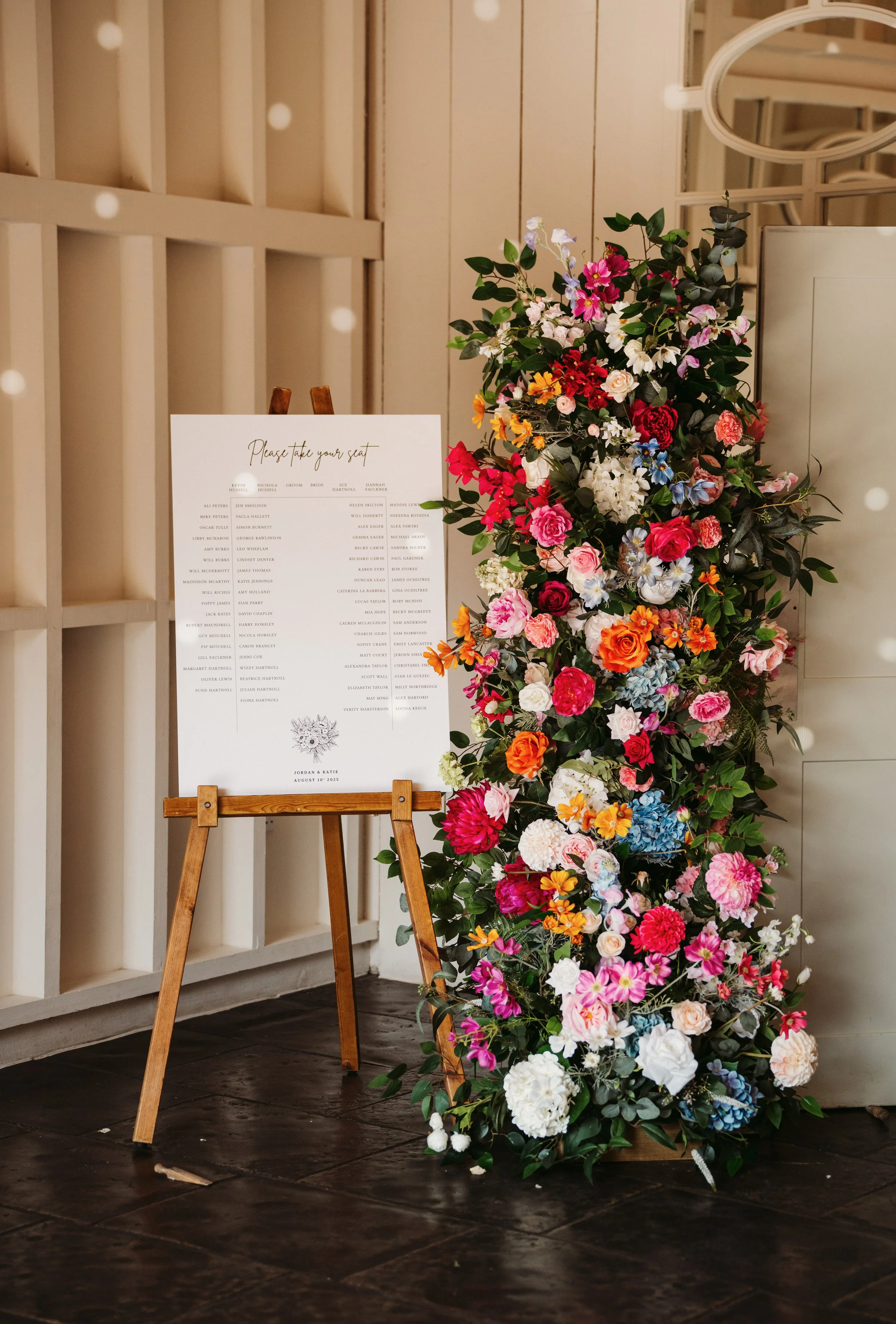 A colorful floral arrangement with white, pink, red, orange, blue, and purple flowers next to a wedding seating chart on a wooden easel.