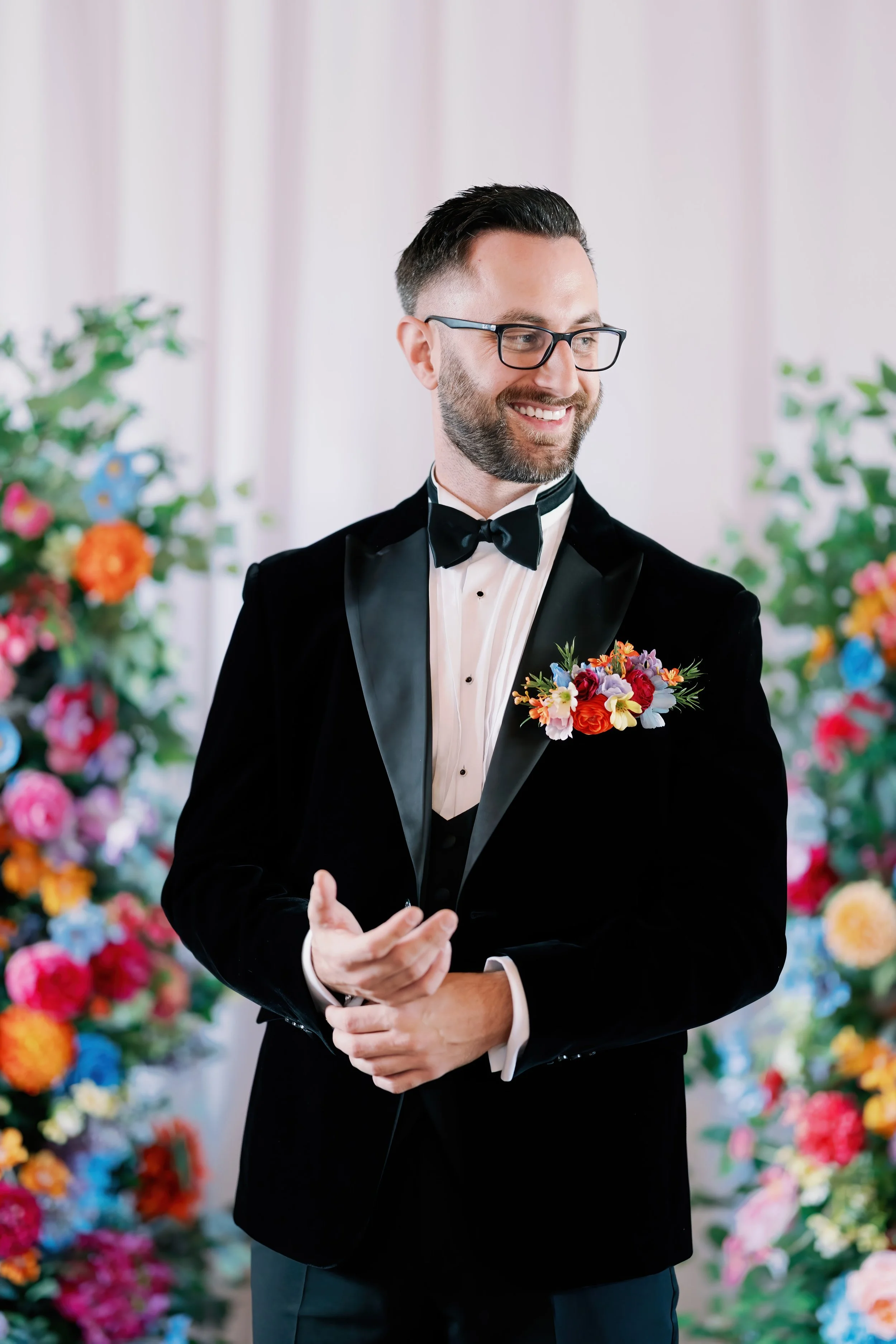 Man in tuxedo with bow tie and glasses, smiling, standing in front of colorful floral arrangements.
