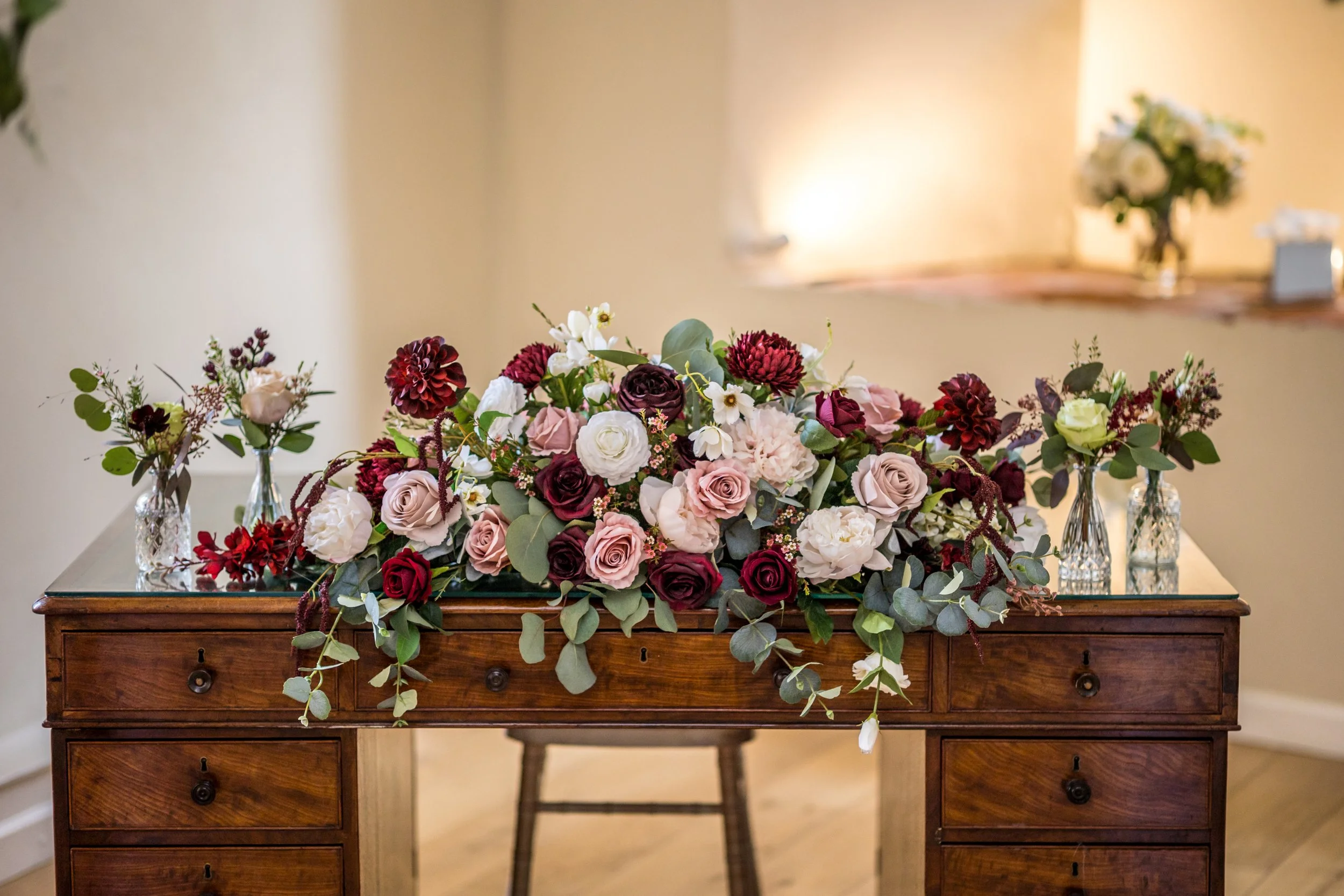 A wooden table decorated with a large arrangement of pink, white, and burgundy roses, surrounded by smaller flower arrangements in glass vases, set indoors.