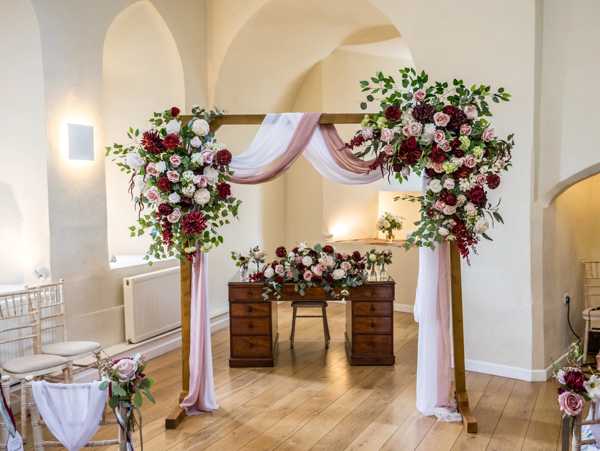 A wedding arch decorated with pink, white, and burgundy flowers and greenery, with pink and white drapes hanging from the top, set up in an indoor venue with beige walls and wooden floors.