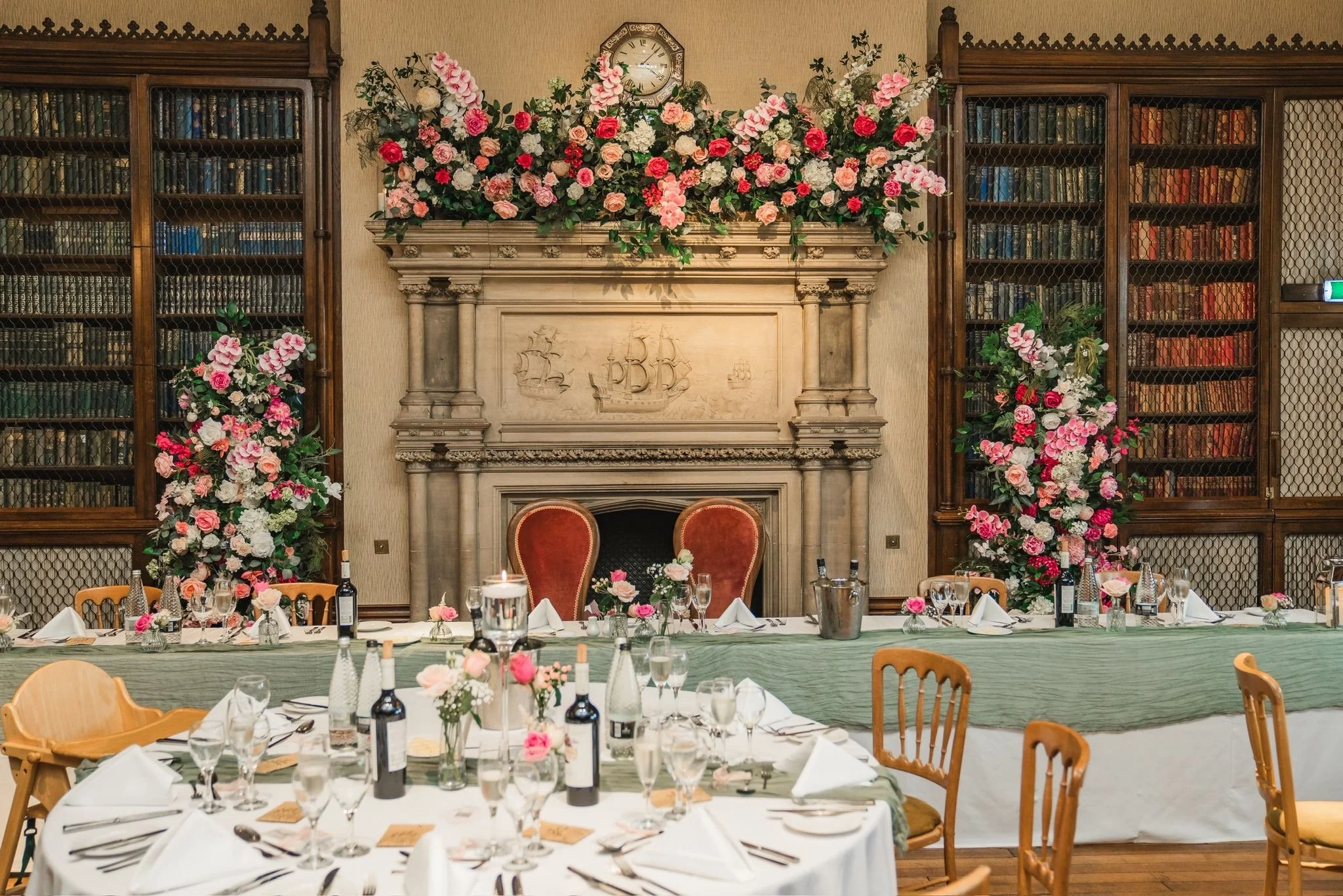 Elegant dining room set for a wedding or special event with arranged tables, wine bottles, glasses, flowers, floral decorations, and a fireplace with an elaborate mantelpiece and floral arrangements, surrounded by bookshelves and wooden chairs.