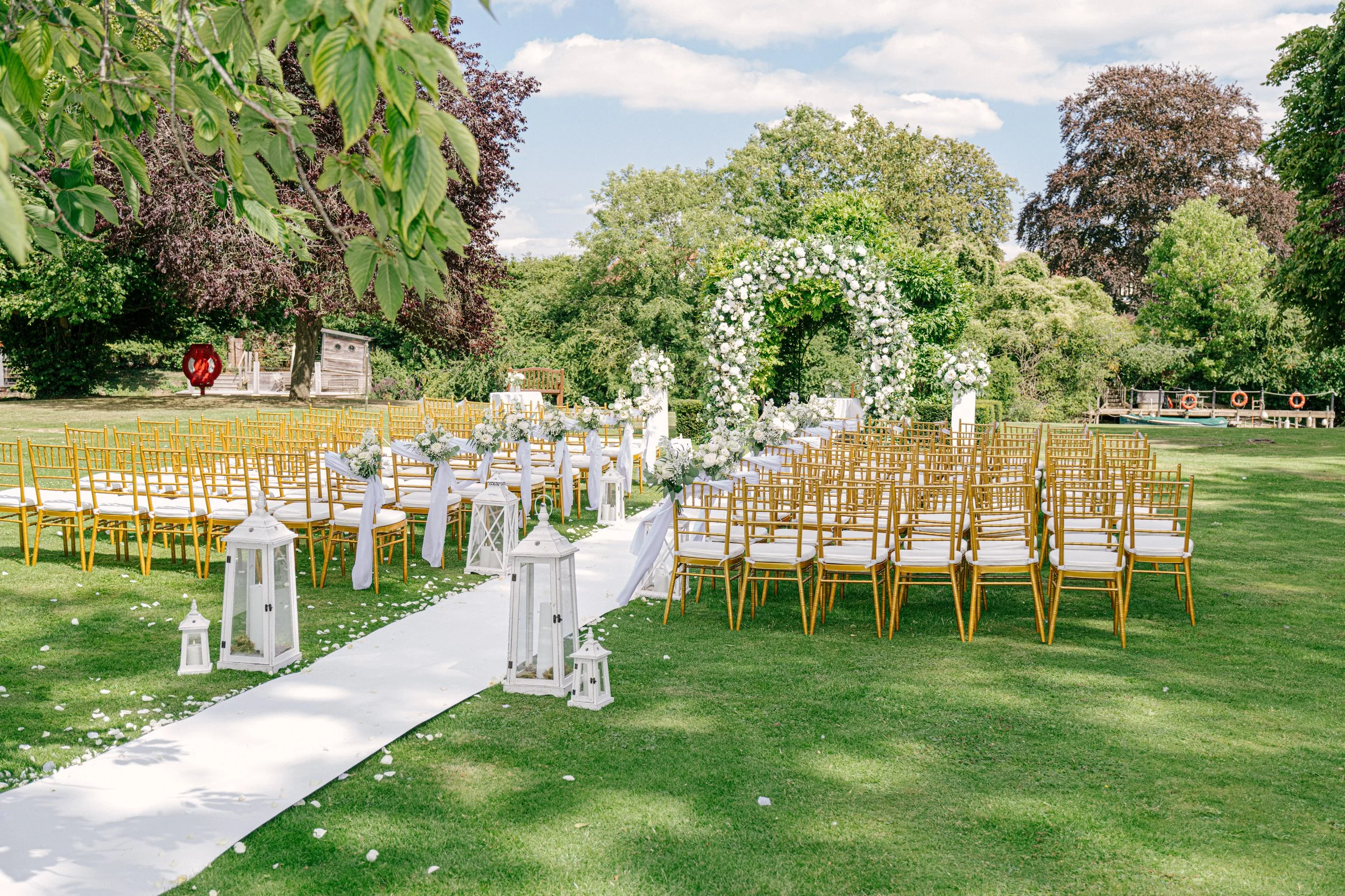 Outdoor wedding setup with rows of gold chairs with white cushions, a floral arch, white lanterns, and a white aisle runner on green grass, surrounded by trees under a partly cloudy sky.