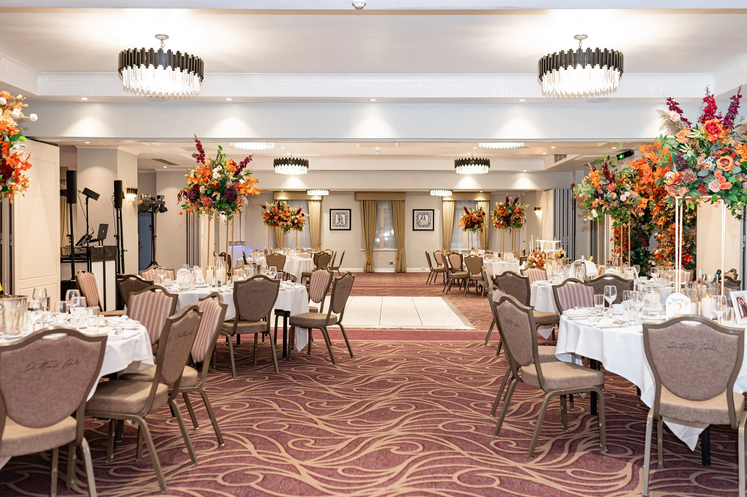 Elegant banquet hall decorated for a wedding reception with round tables covered in white tablecloths, floral centerpieces, and chairs with beige upholstery. There is a small dance floor in the center, chandeliers overhead, and large floral arrangeme