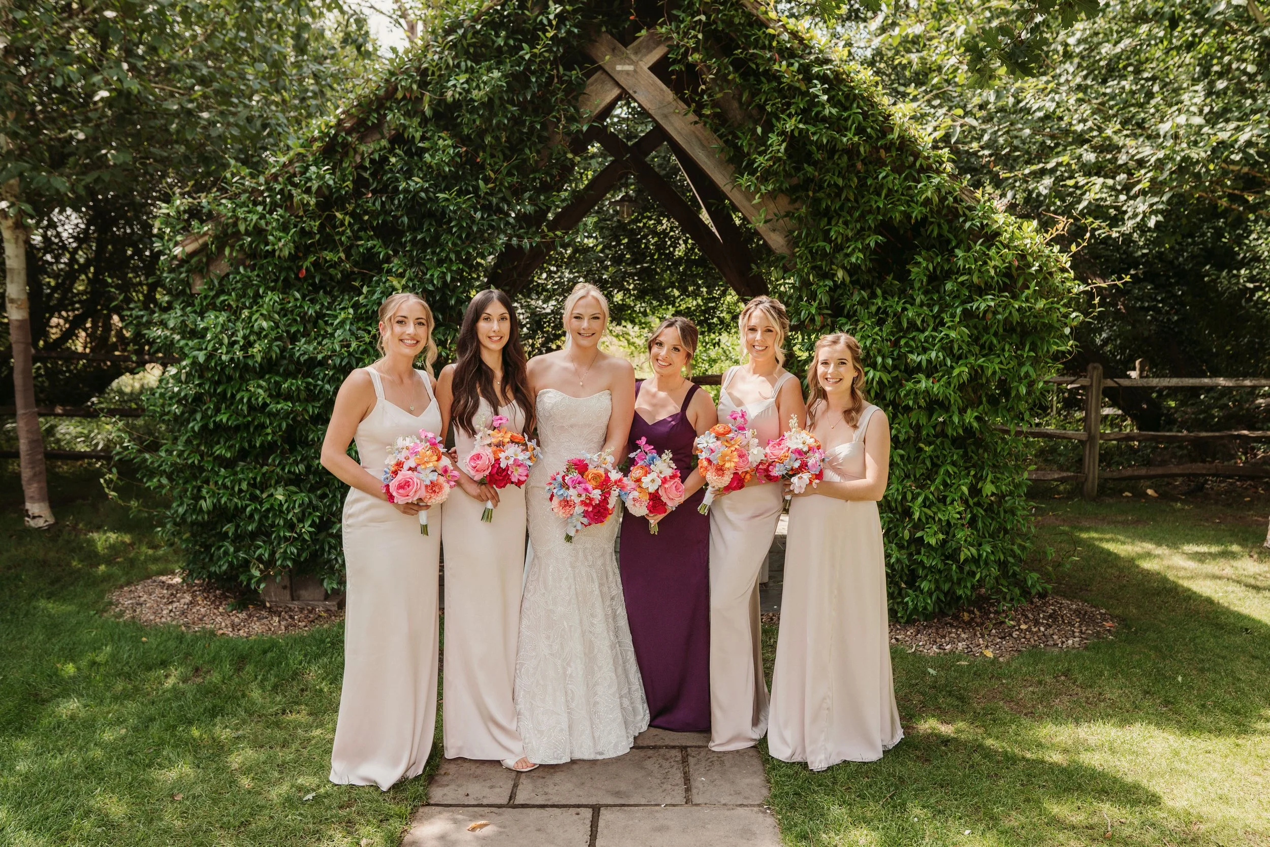 A group of six women, including a bride in a white wedding dress, holding bouquets of pink, orange, and purple flowers, standing in front of a lush green archway outdoors during daytime.