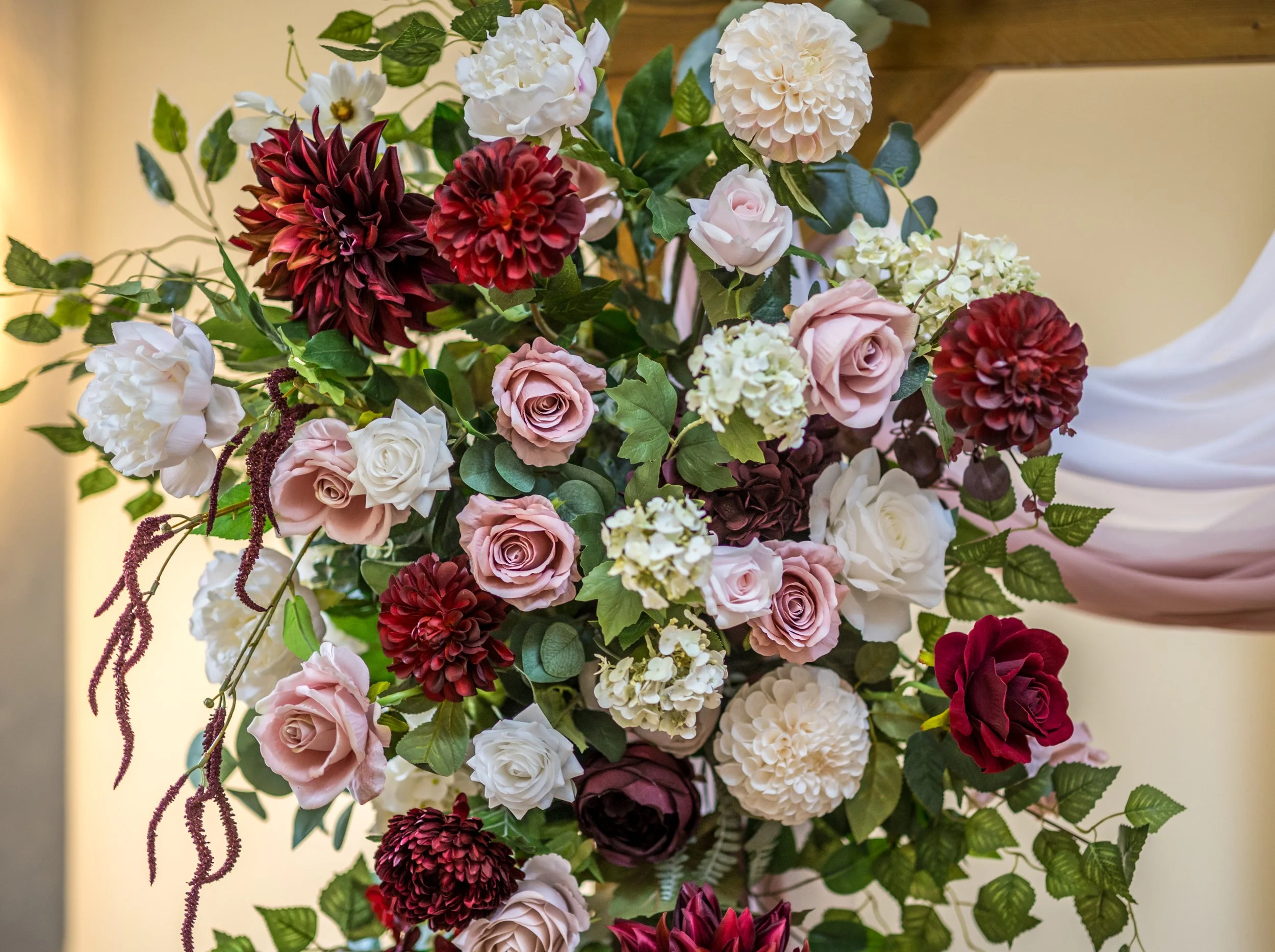 A bouquet of flowers with pink, white, and dark red roses, white hydrangeas, and burgundy dahlias, surrounded by green leaves and vines.