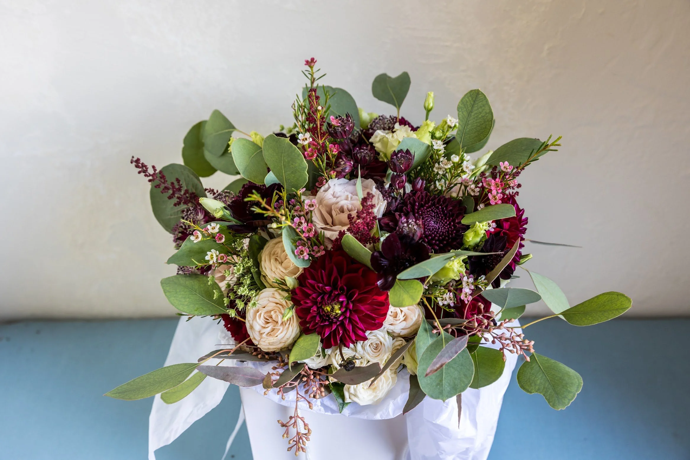 Arranged bouquet of various flowers including roses, dahlias, and small pink blossoms with green leaves in white wrapping paper.