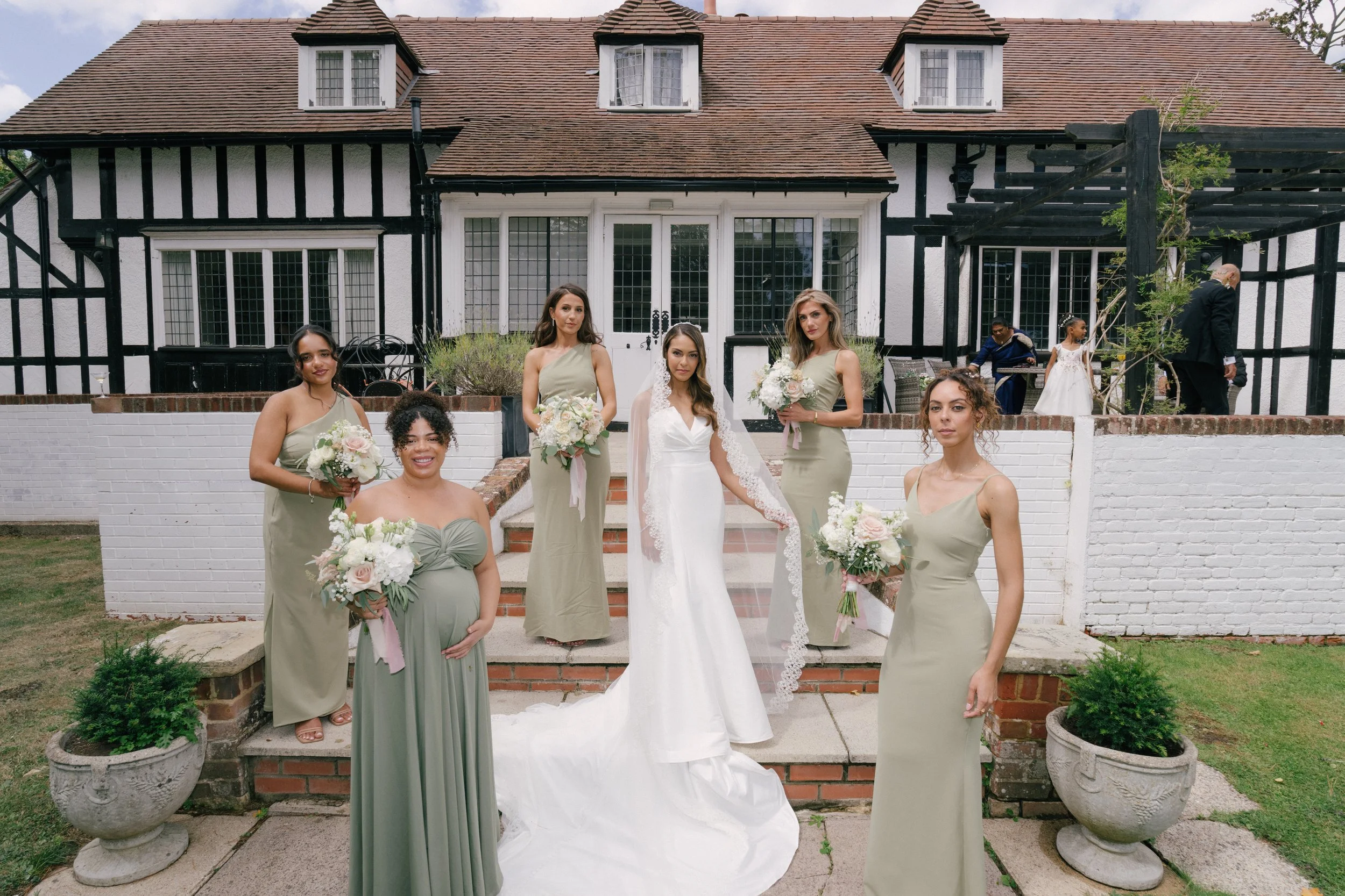 Wedding party standing on steps outside a black and white timber-framed house. The bride in a white gown with lace veil, surrounded by bridesmaids in sage green dresses, holding bouquets of white and pink flowers.